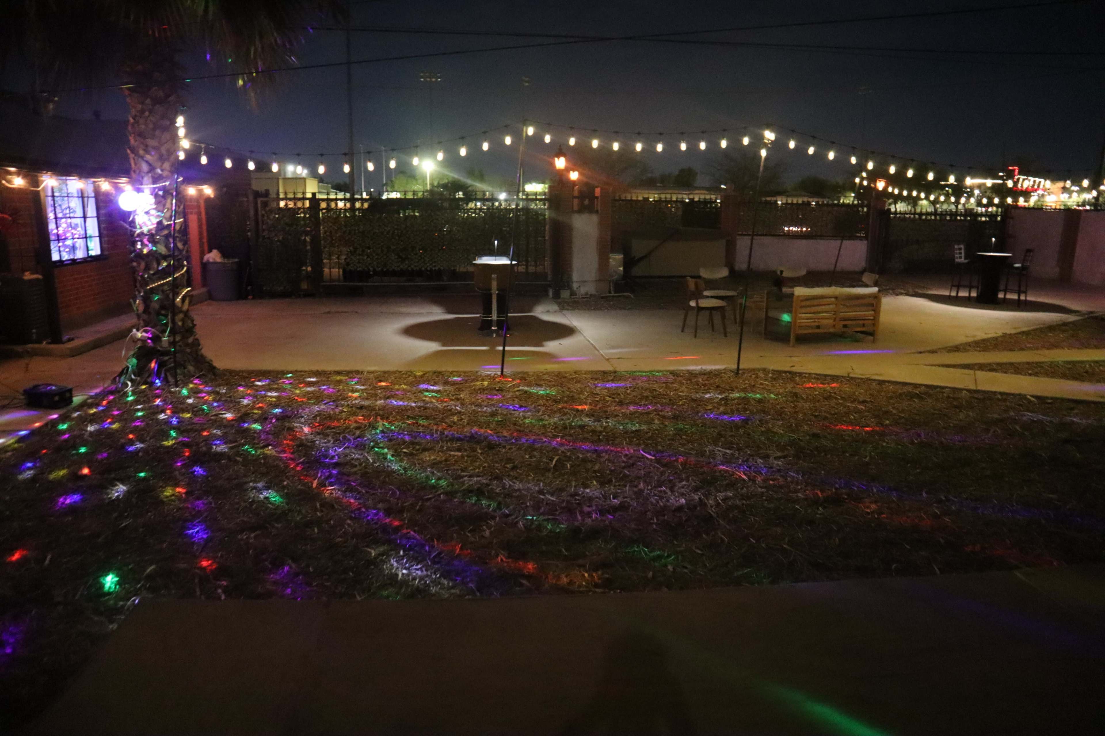 The image shows an outdoor space at night with colorful lights projected onto the ground and string lights hung above, creating a festive atmosphere.