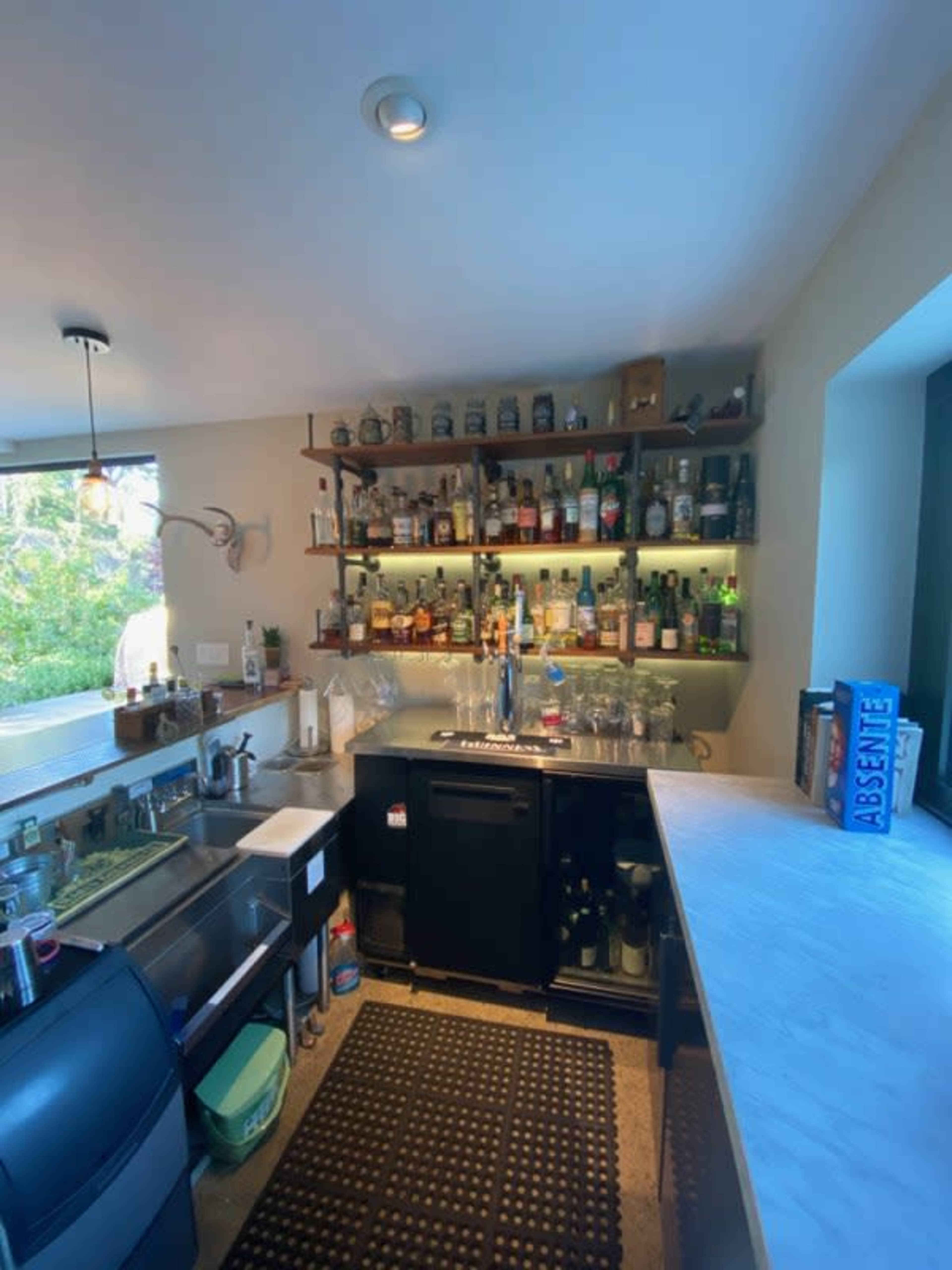 A well-organized home bar area featuring a variety of bottles on shelves, a sink, a cooler, and a marble countertop.
