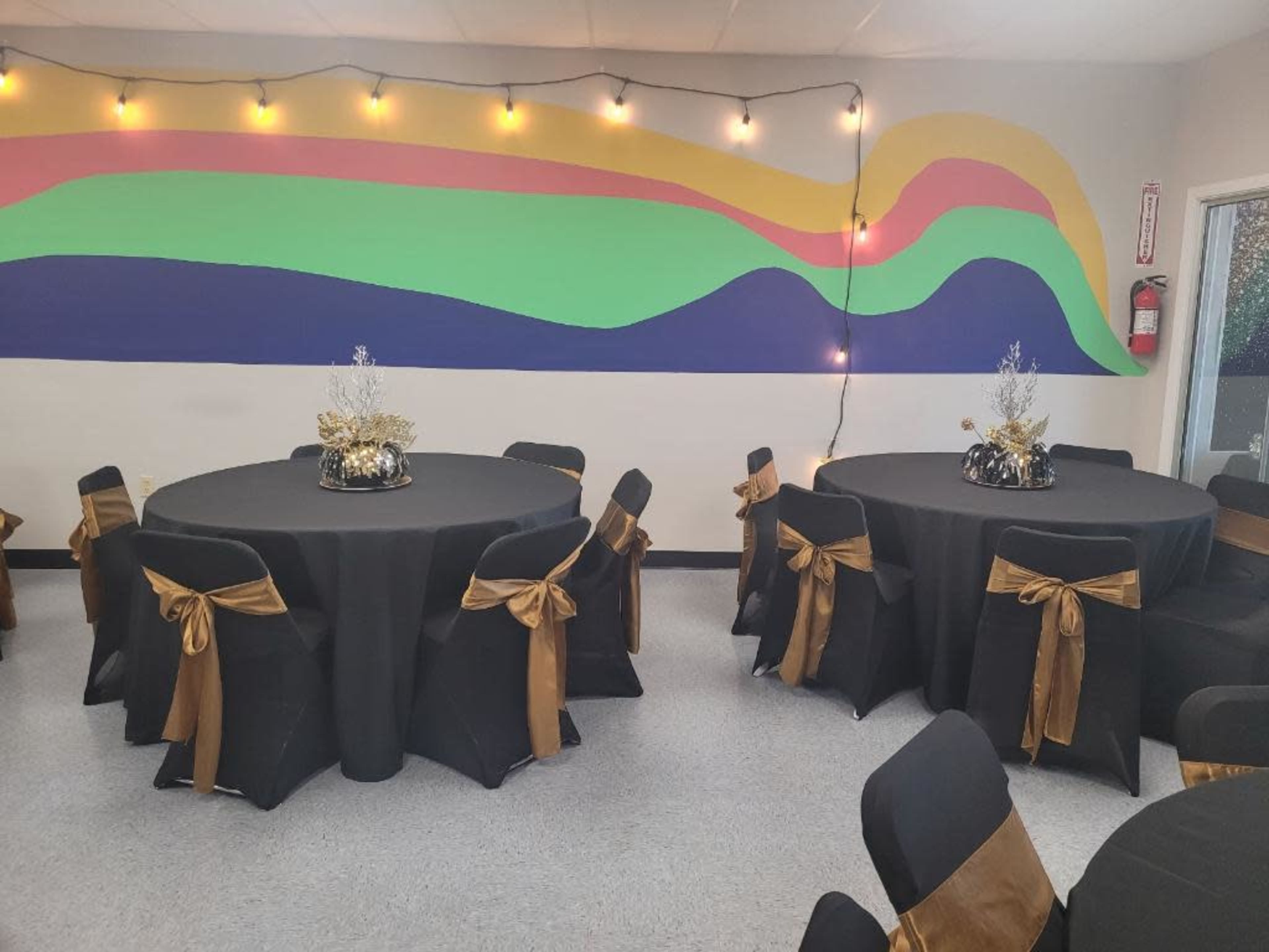 The image shows a decorated indoor event space with two round tables covered in black tablecloths and gold bows on black chairs, set against a colorful mural and string lights.
