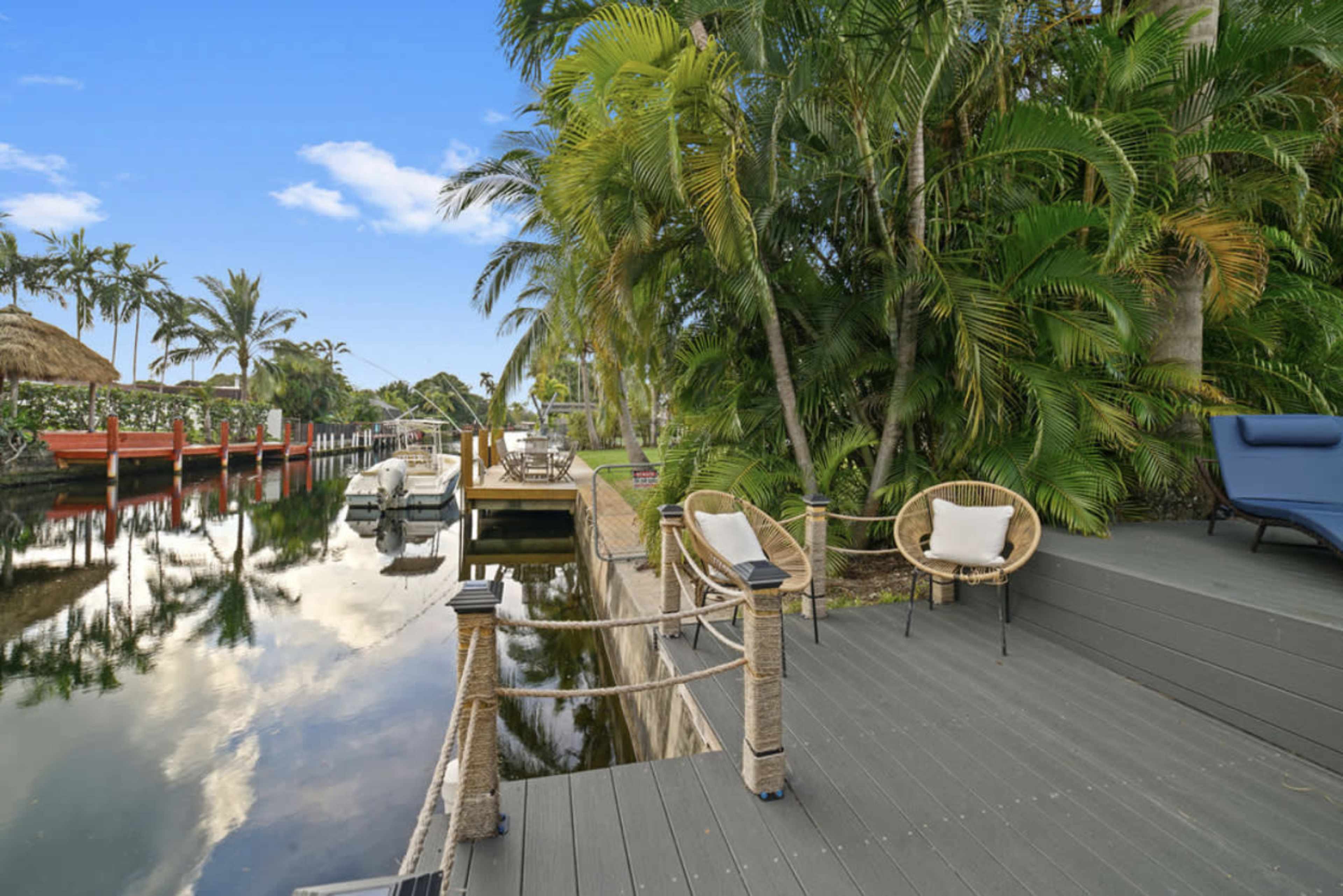 A dock along a calm waterway is lined with greenery, featuring a pair of chairs and a boat moored nearby.