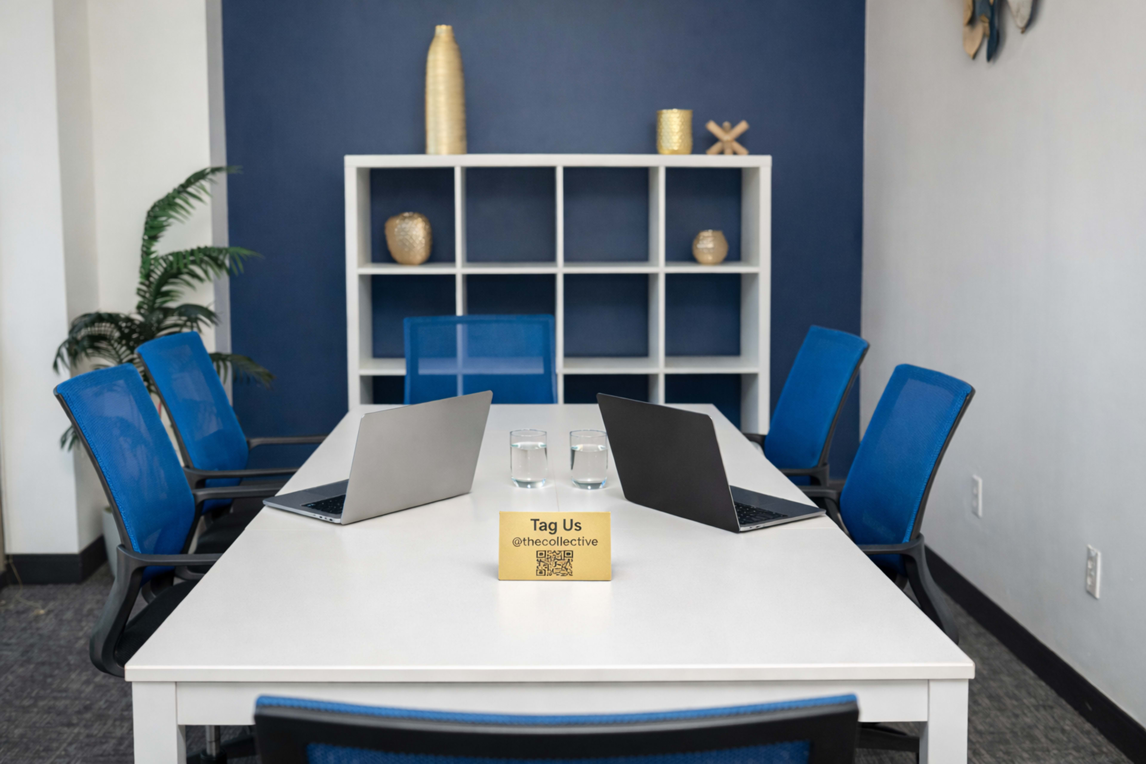 A modern conference room features a white table with two laptops open, surrounded by six blue chairs, and a decorative shelf against a blue wall.