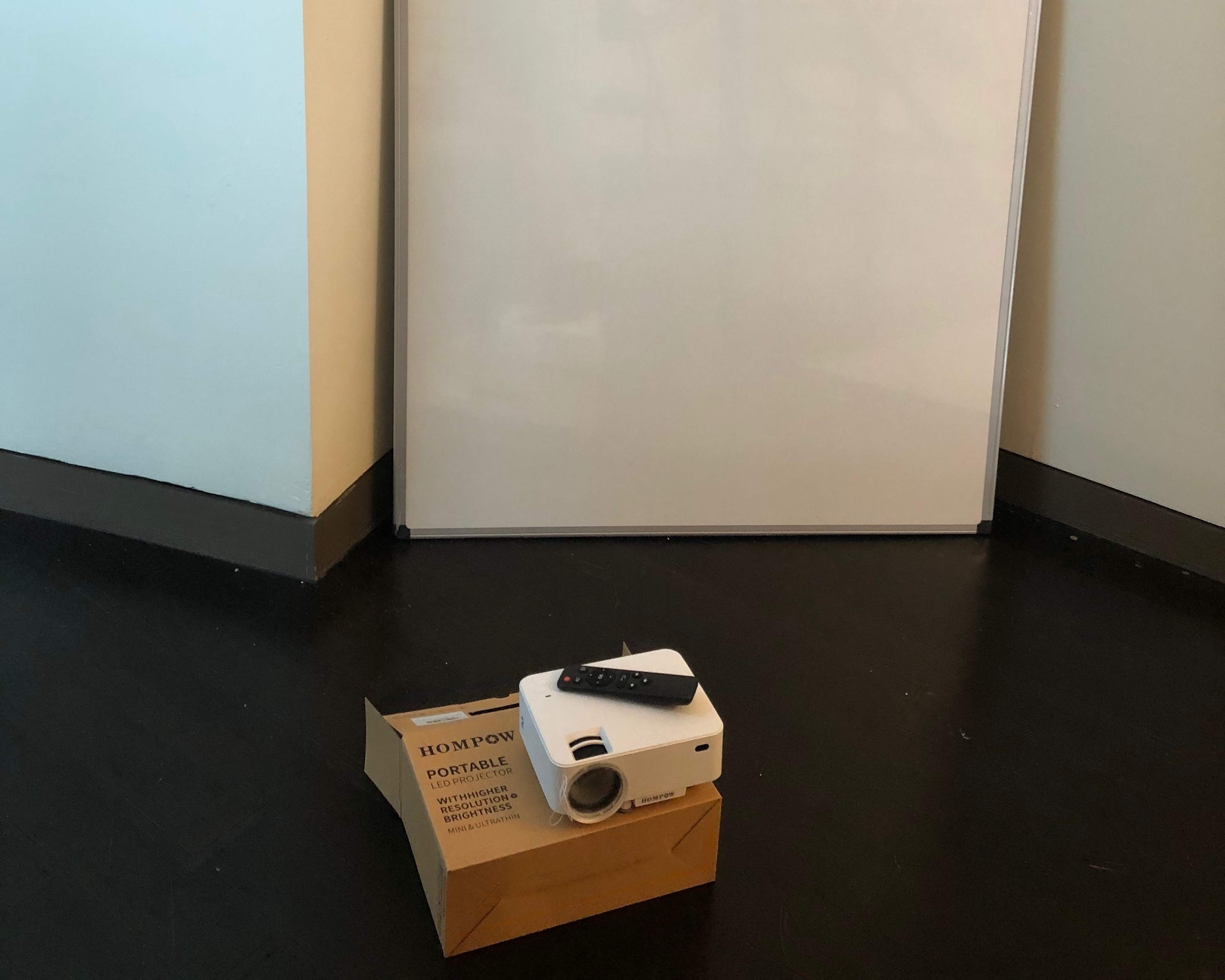A small projector sits on a cardboard box in front of a blank white projection screen.