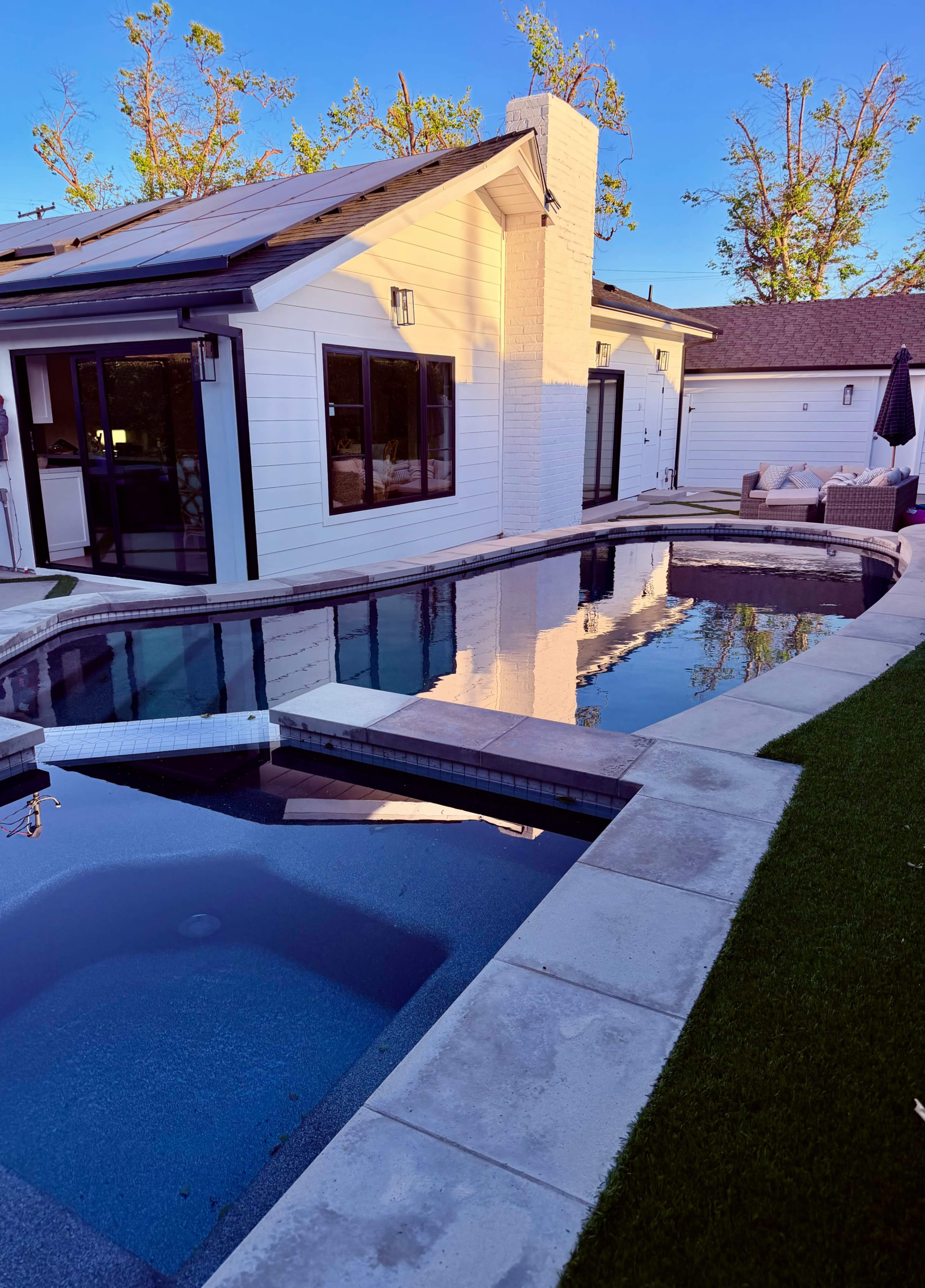 The image shows a modern backyard featuring a swimming pool with a hot tub, surrounded by landscaped greenery and a house with large windows.