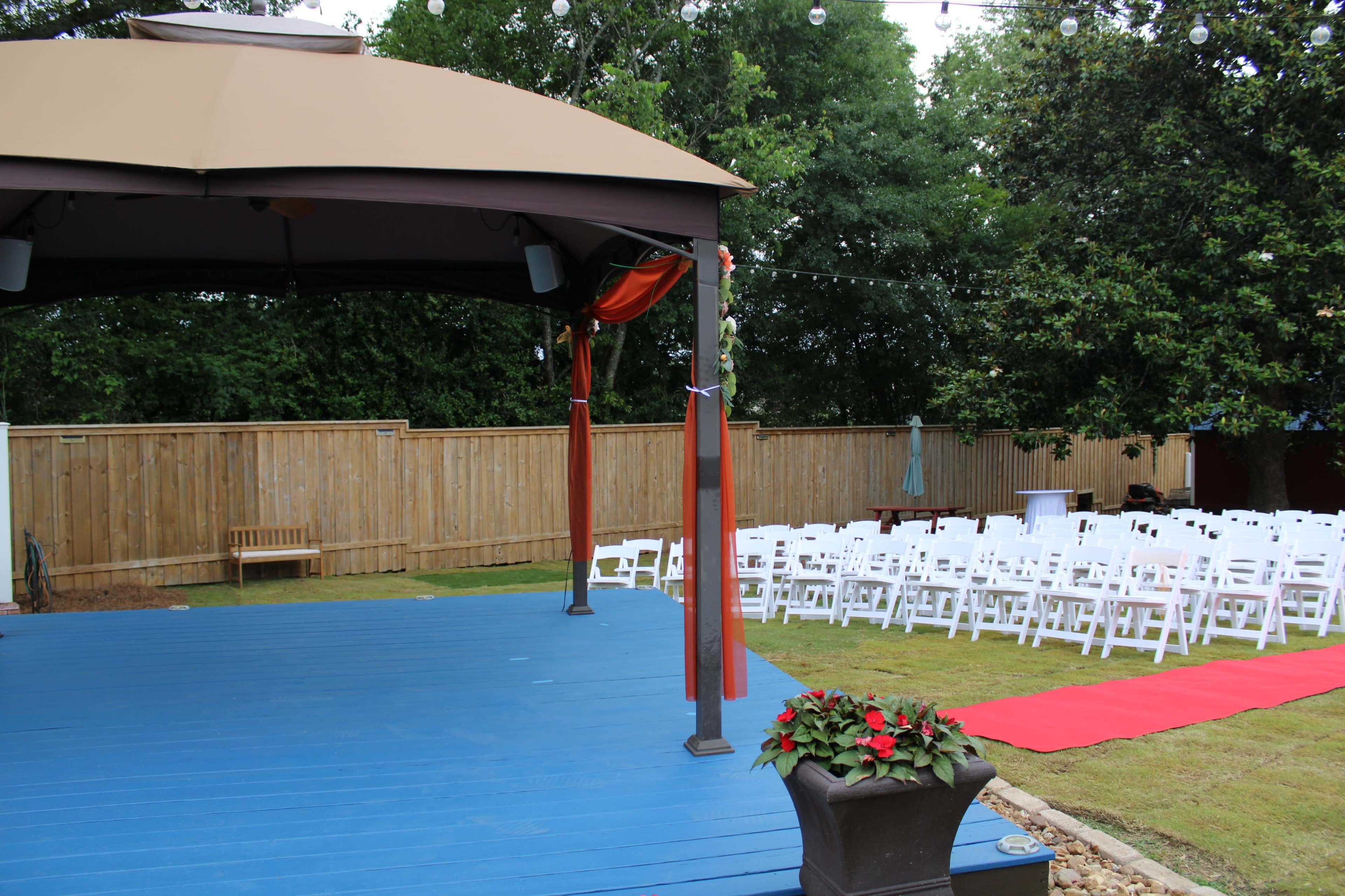 A blue stage with a canopy is set up in a garden facing rows of white chairs arranged on green grass, with a red carpet leading towards the stage.