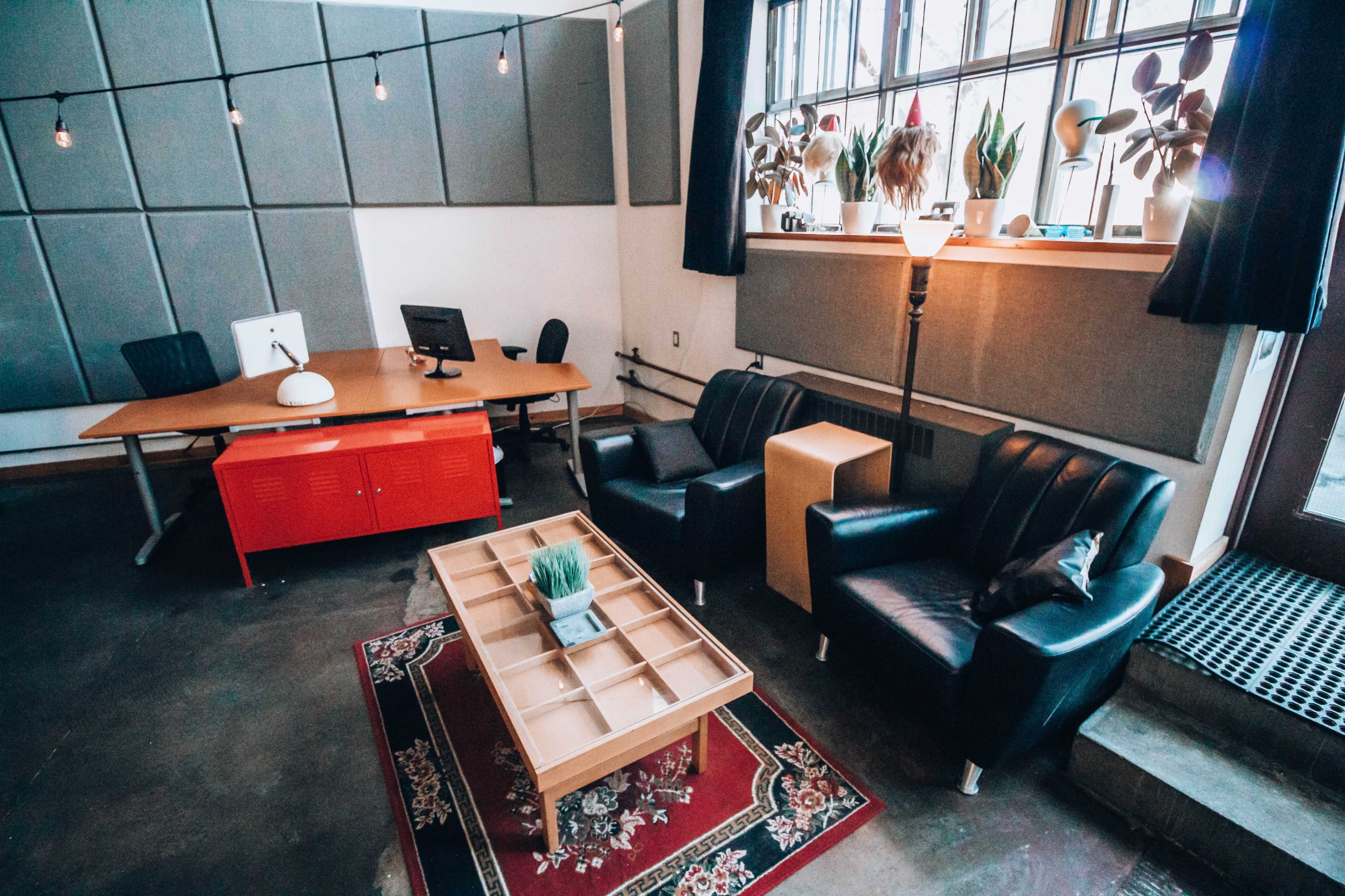 A modern office space with a wooden desk, black chairs, a coffee table, and large windows adorned with plants.