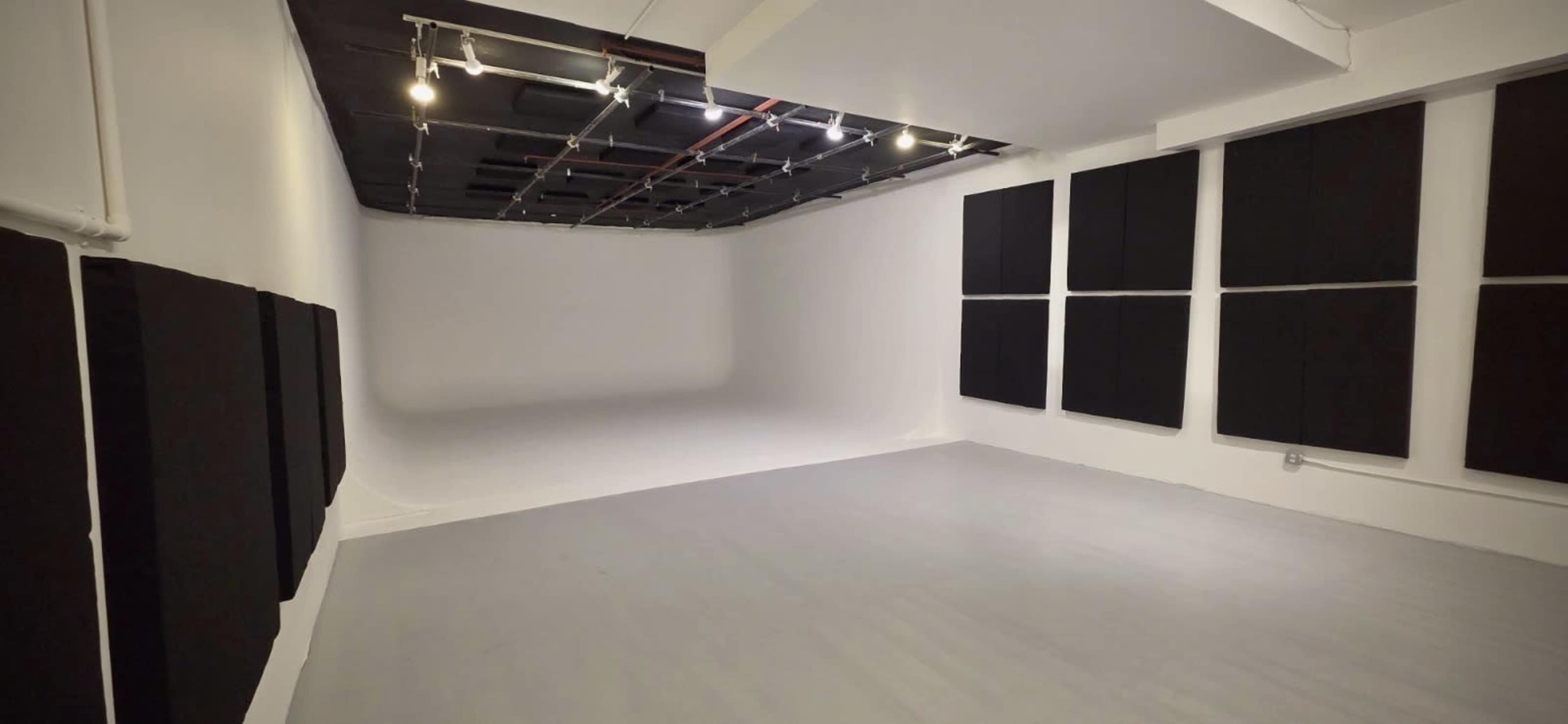 The image shows a spacious, empty studio with white walls and several black soundproofing panels mounted on the walls.