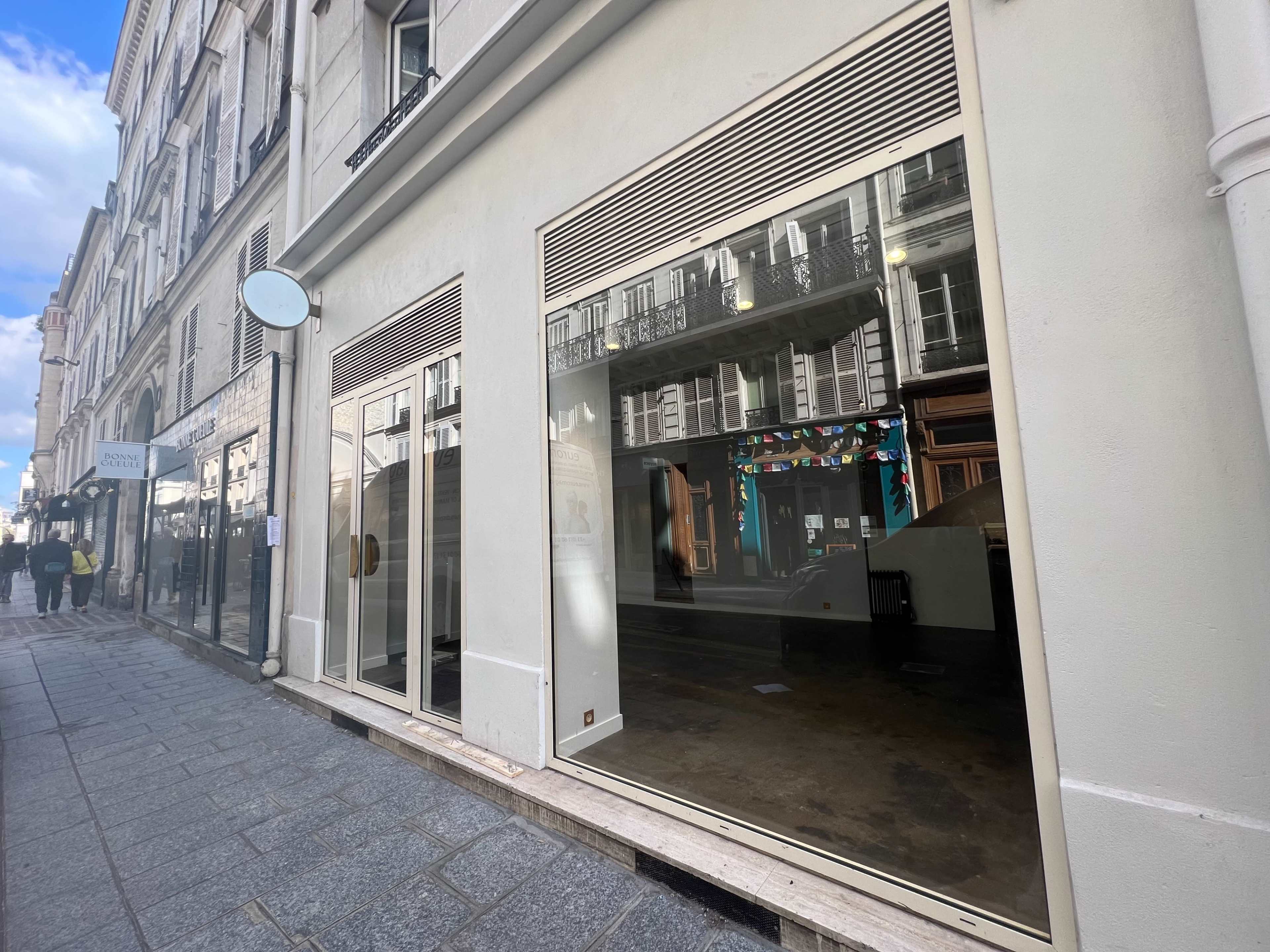 The image shows a vacant storefront with large glass windows on a sidewalk in a city.