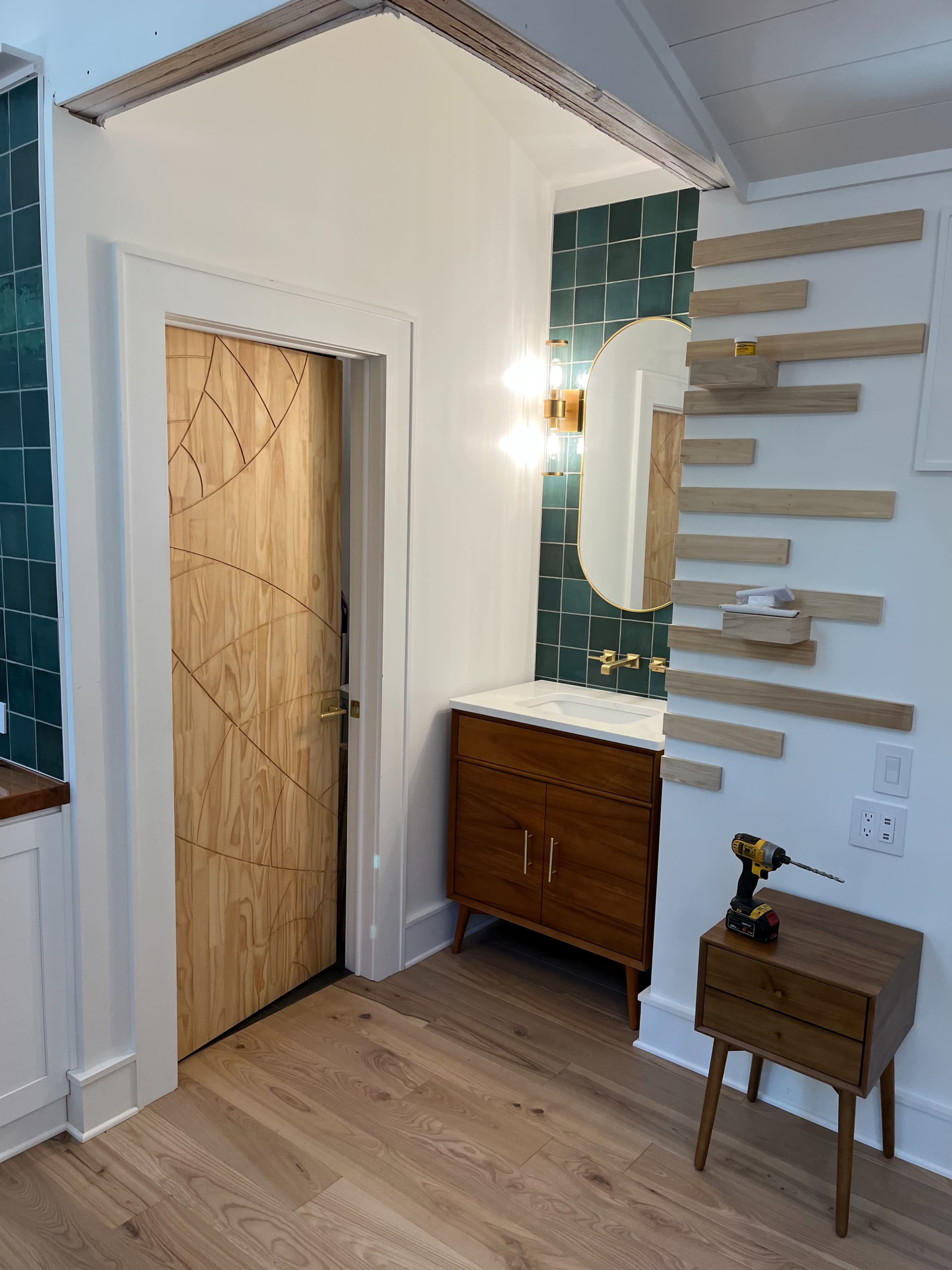 The image shows a modern interior featuring a wooden door with a patterned design, a mirror above a sink with a countertop, and a series of wooden shelves mounted on the wall.