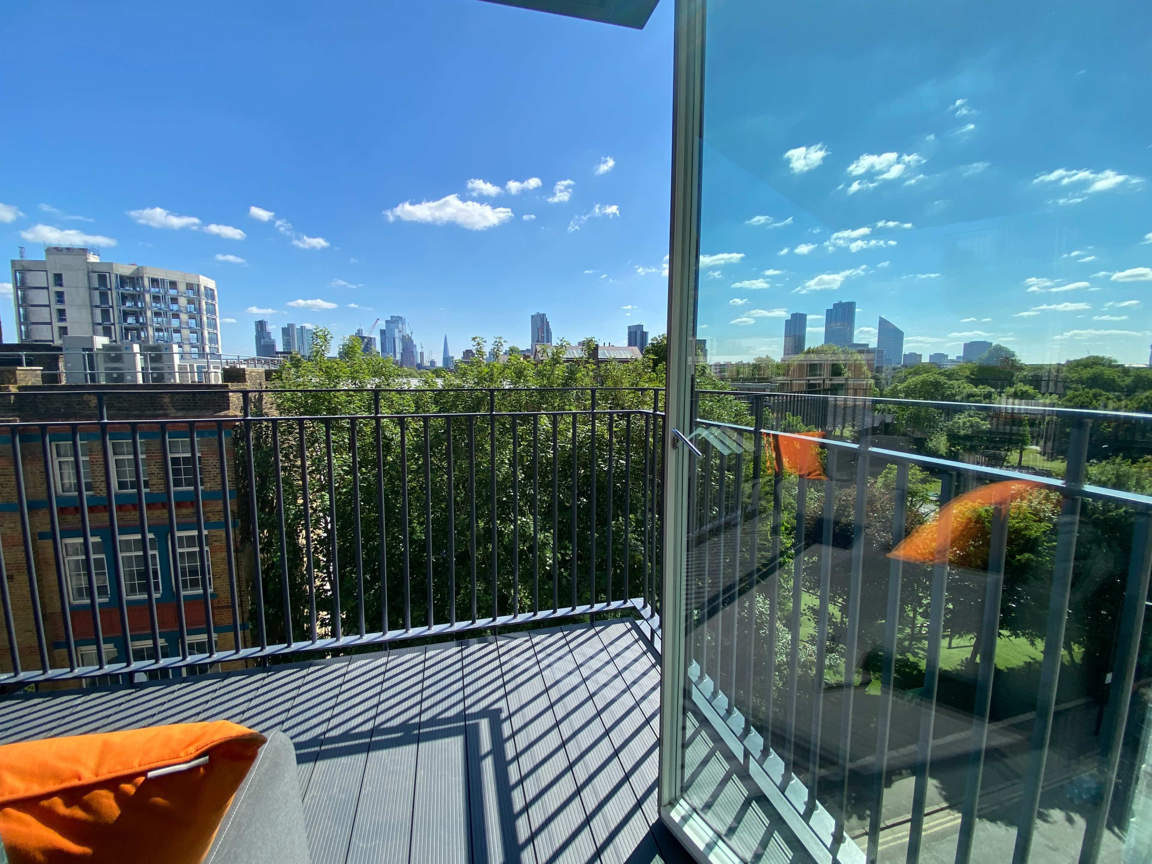A balcony with a clear glass railing overlooks a city skyline and green trees under a blue sky filled with scattered clouds.
