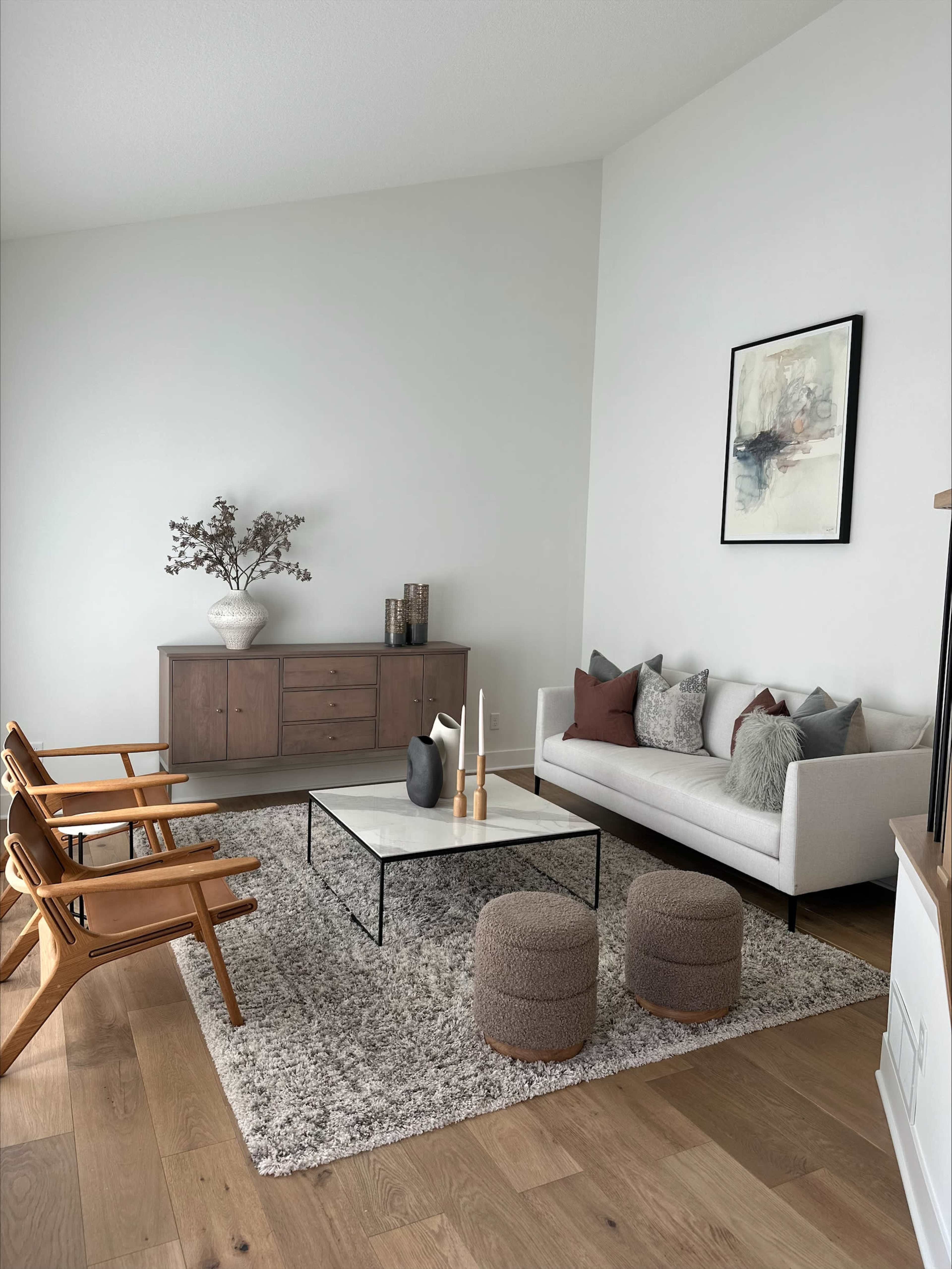 A modern living room featuring a light-colored wall, a gray rug, a coffee table, a couch with decorative pillows, a wooden sideboard, and three wooden chairs.