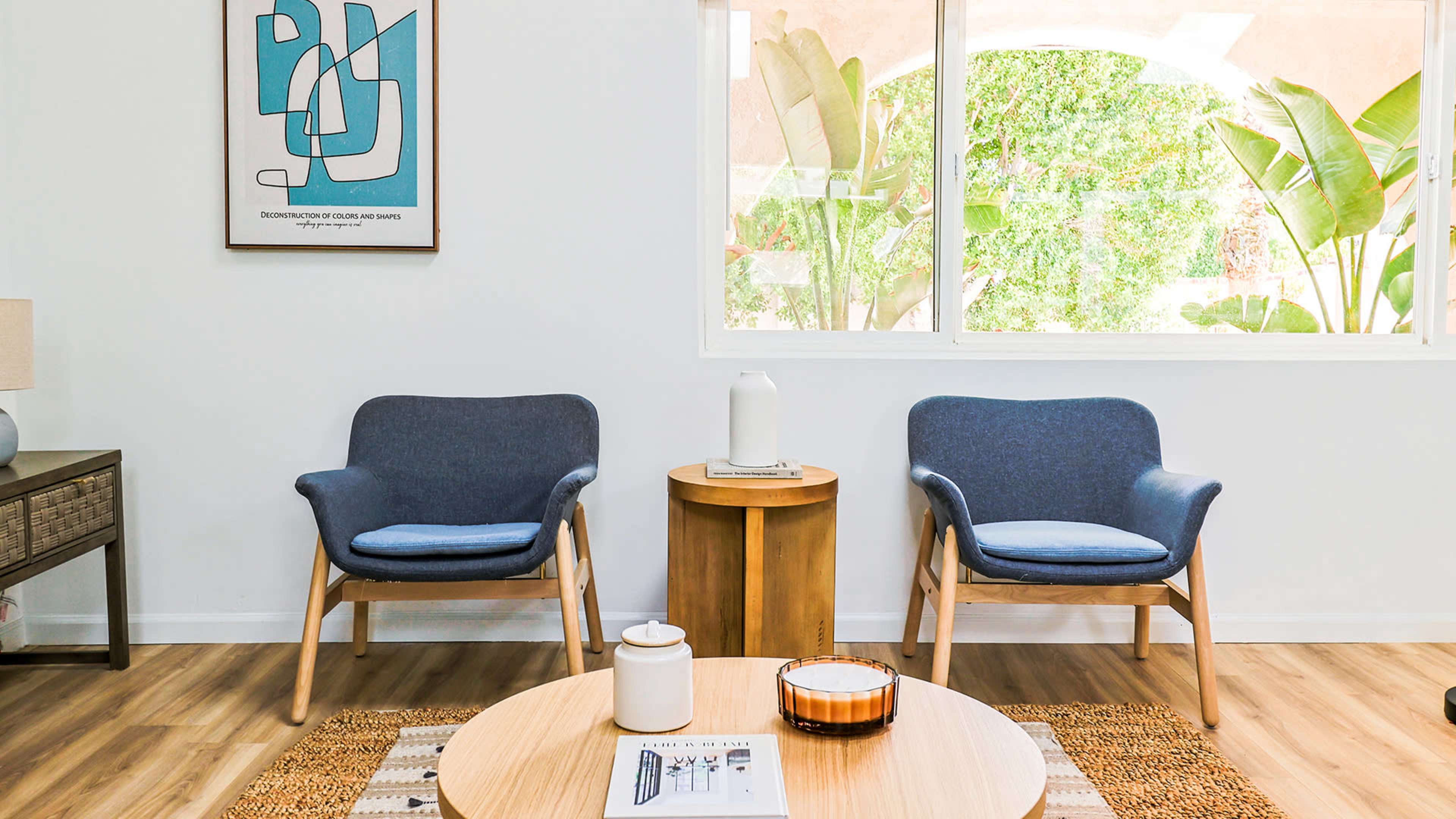 A minimalist living area featuring two blue armchairs, a wooden coffee table, and a light-colored rug, with greenery visible through a large window.