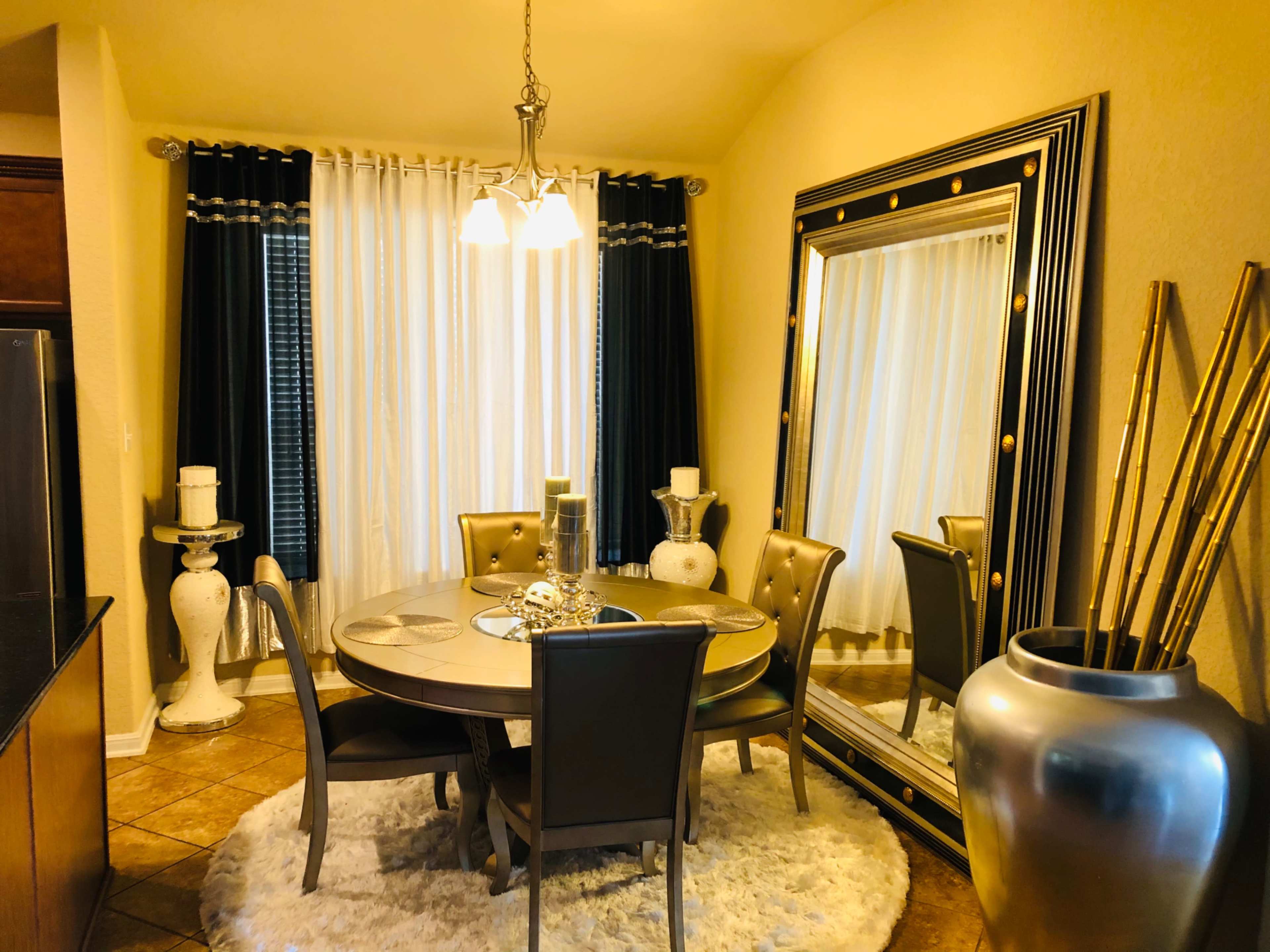 A dining area features a round table surrounded by upholstered chairs, a large decorative mirror, and tall vases against a backdrop of sheer and dark curtains.