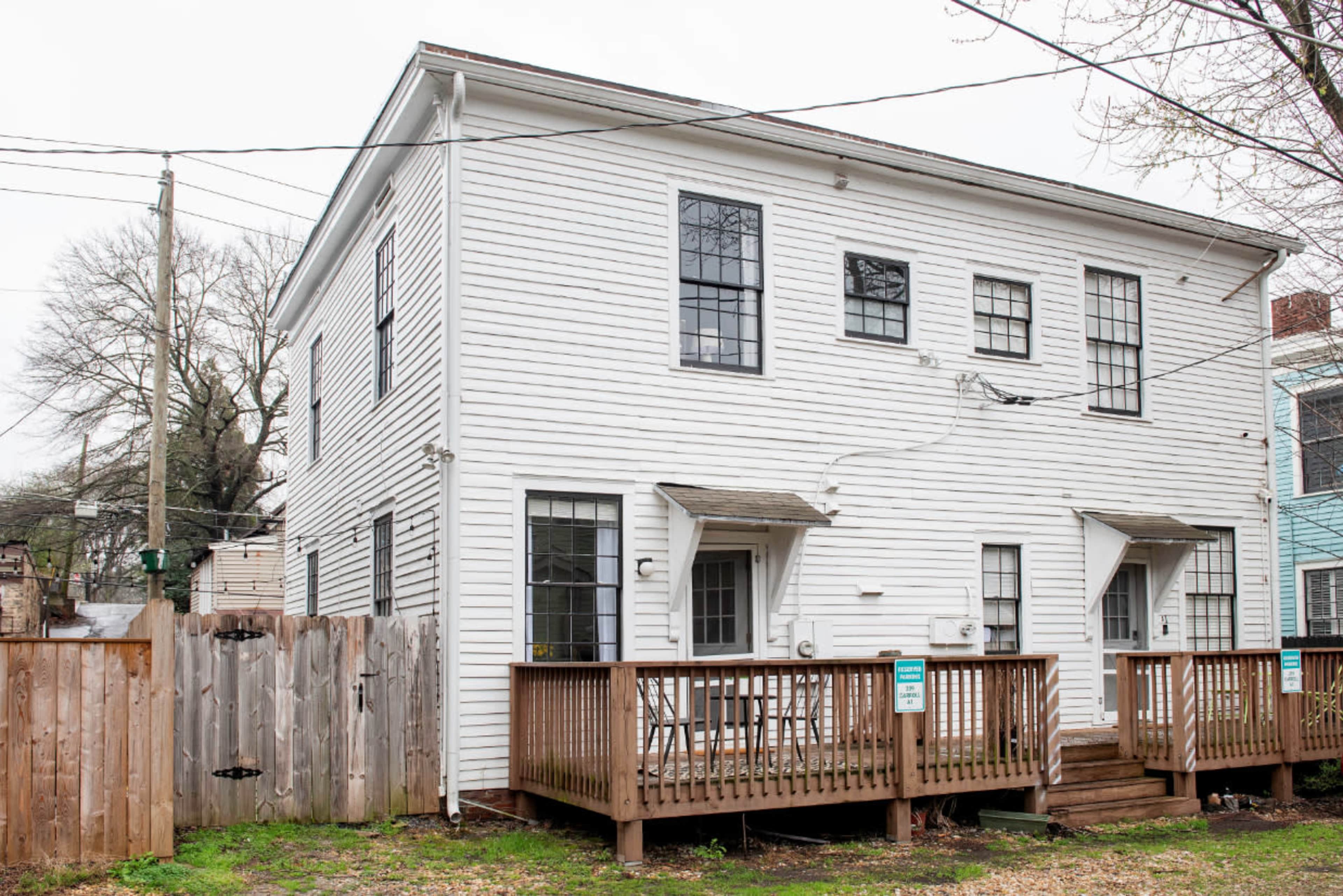 A white two-story house with a wooden deck and two entrances is situated next to a wooden fence.