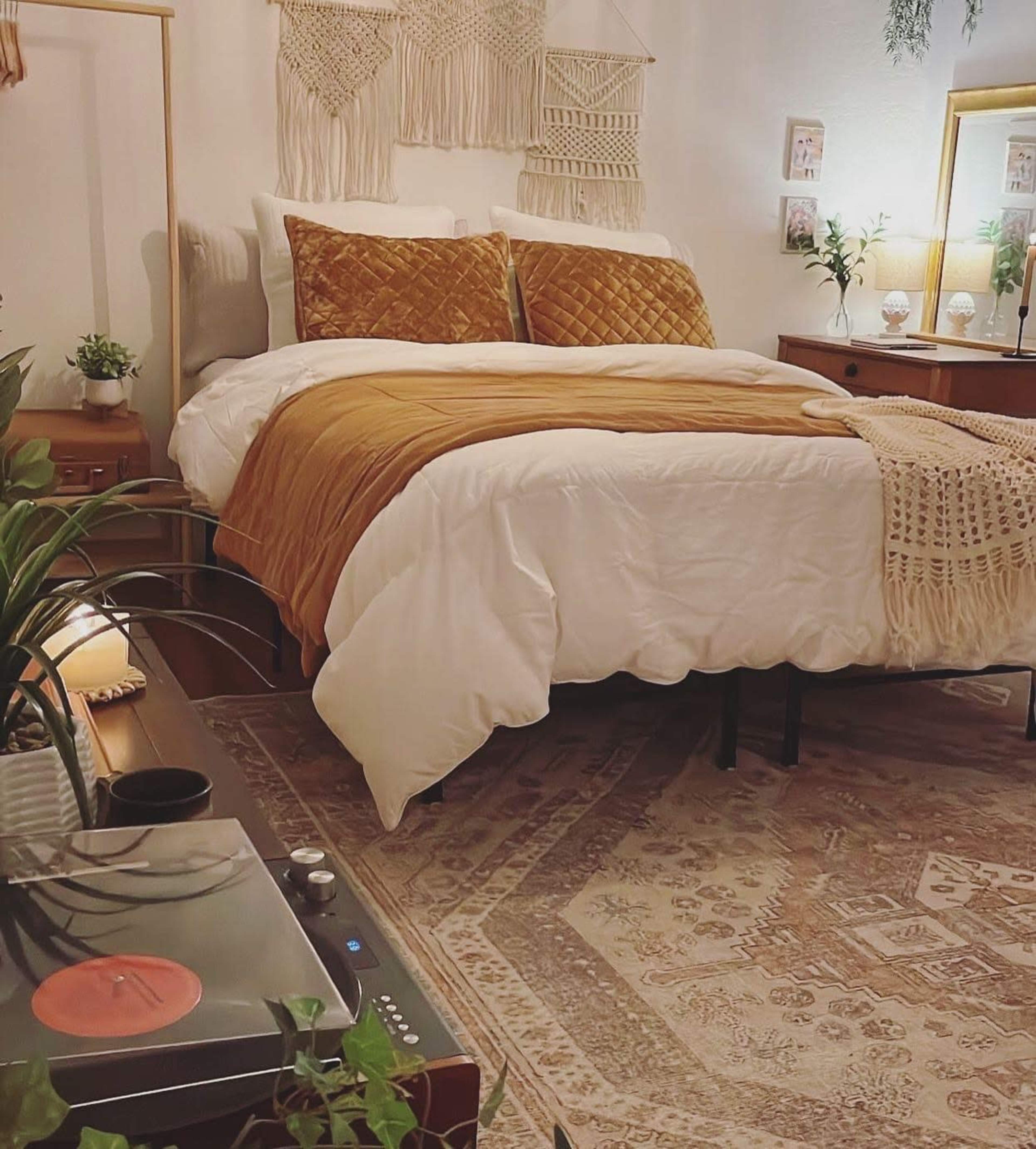 The image shows a cozy bedroom featuring a bed with a white and gold quilt, a patterned rug, and plants arranged around the room.