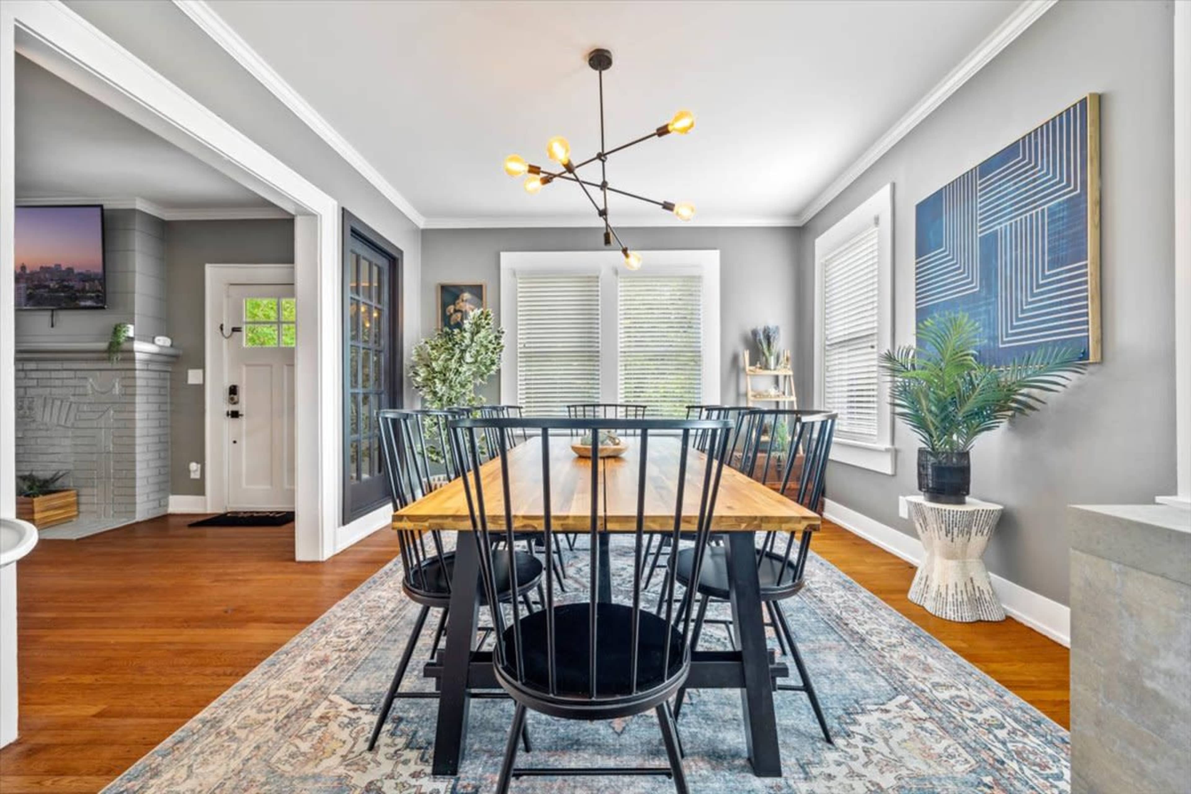 A wooden dining table surrounded by black chairs is set in a bright room with gray walls and decorative plants.