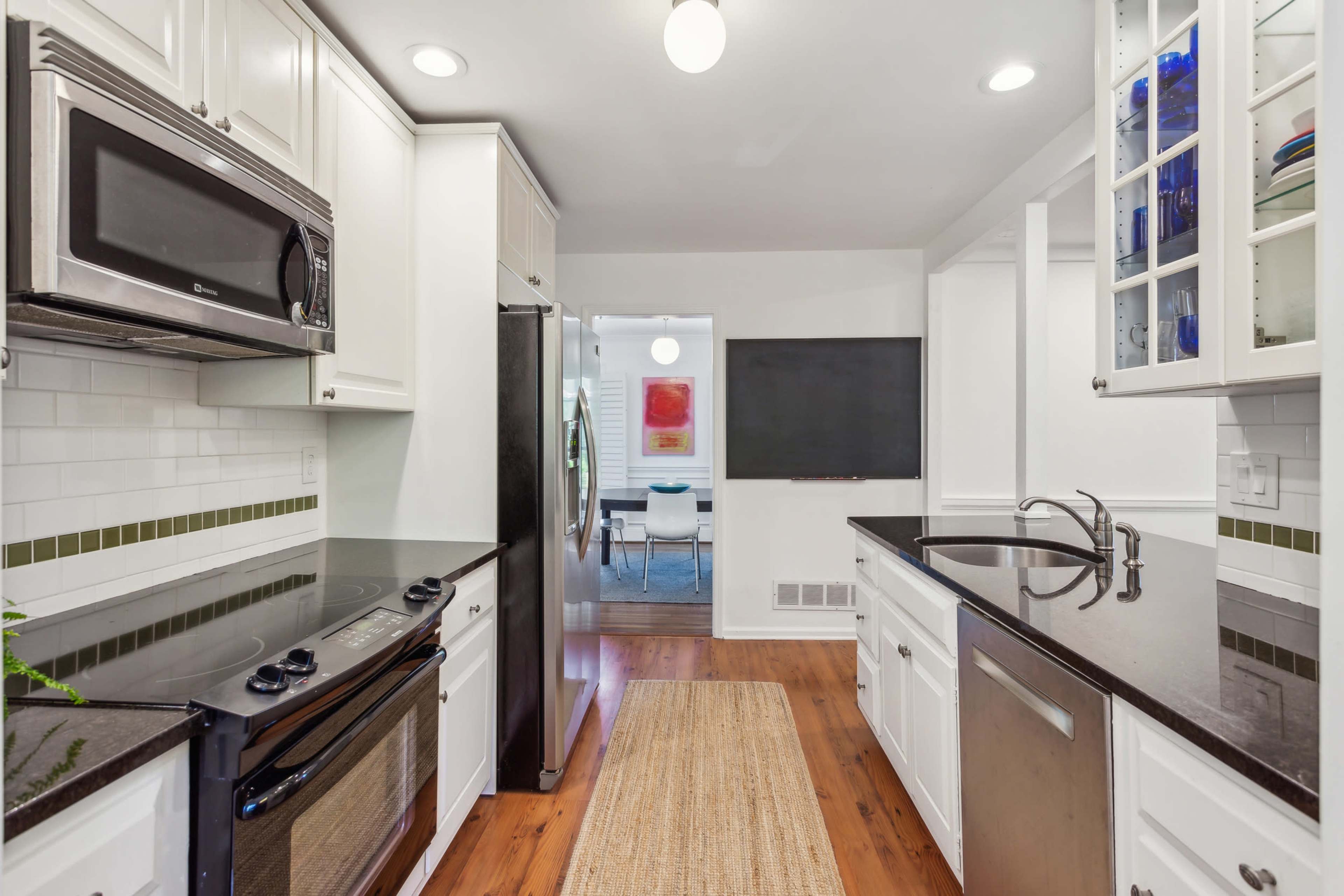 The image shows a modern kitchen with white cabinets, black countertops, and stainless steel appliances, leading to a dining area in the background.