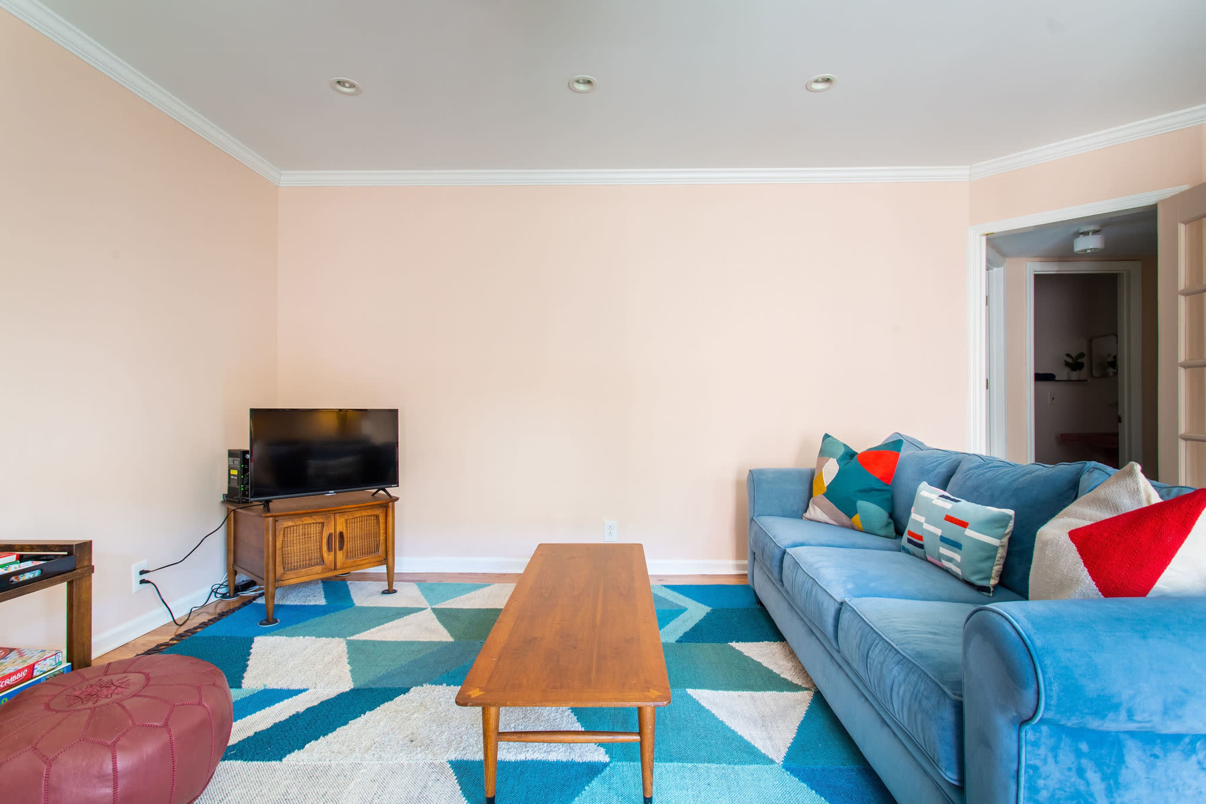 A living room features a blue sofa with colorful pillows, a wooden coffee table, a television, and a geometric-patterned rug on a light-colored floor.