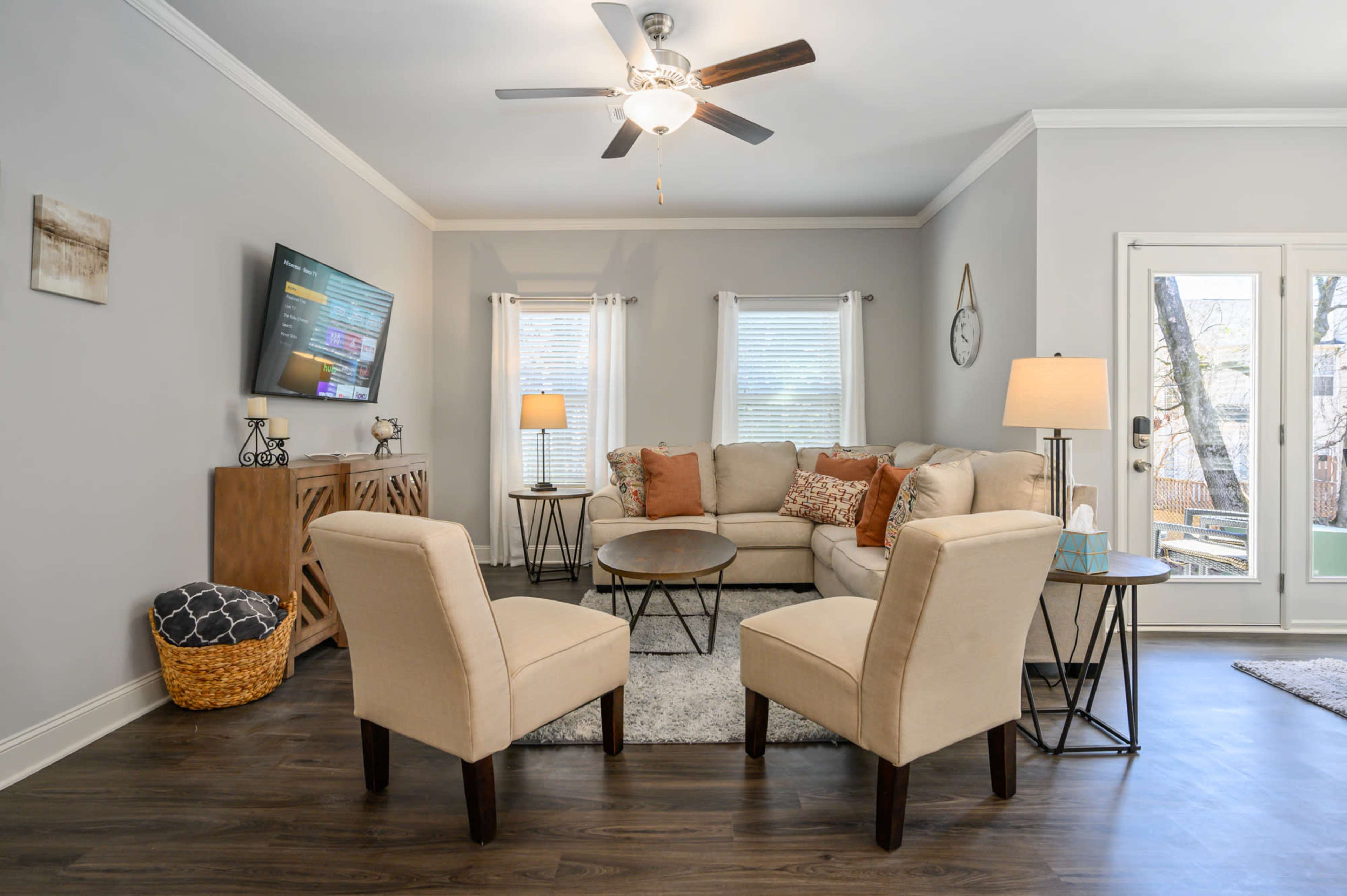 A cozy living room with a light-colored sofa, two chairs, a coffee table, a television mounted on the wall, and large windows that open to a view of the outdoors.