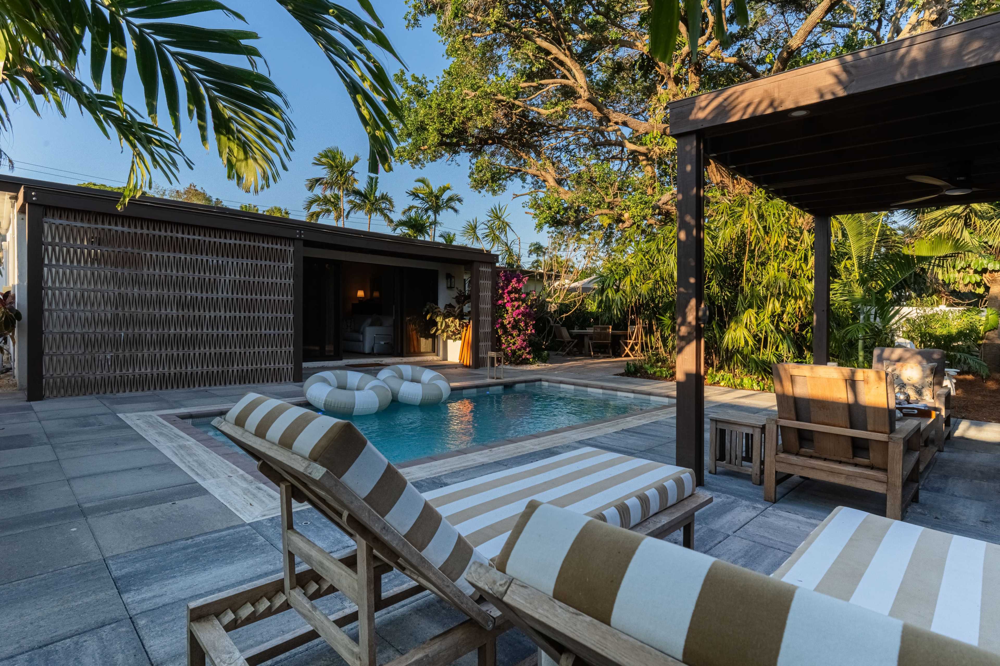 A bright backyard features a pool surrounded by lounge chairs, tropical plants, and a shaded seating area.