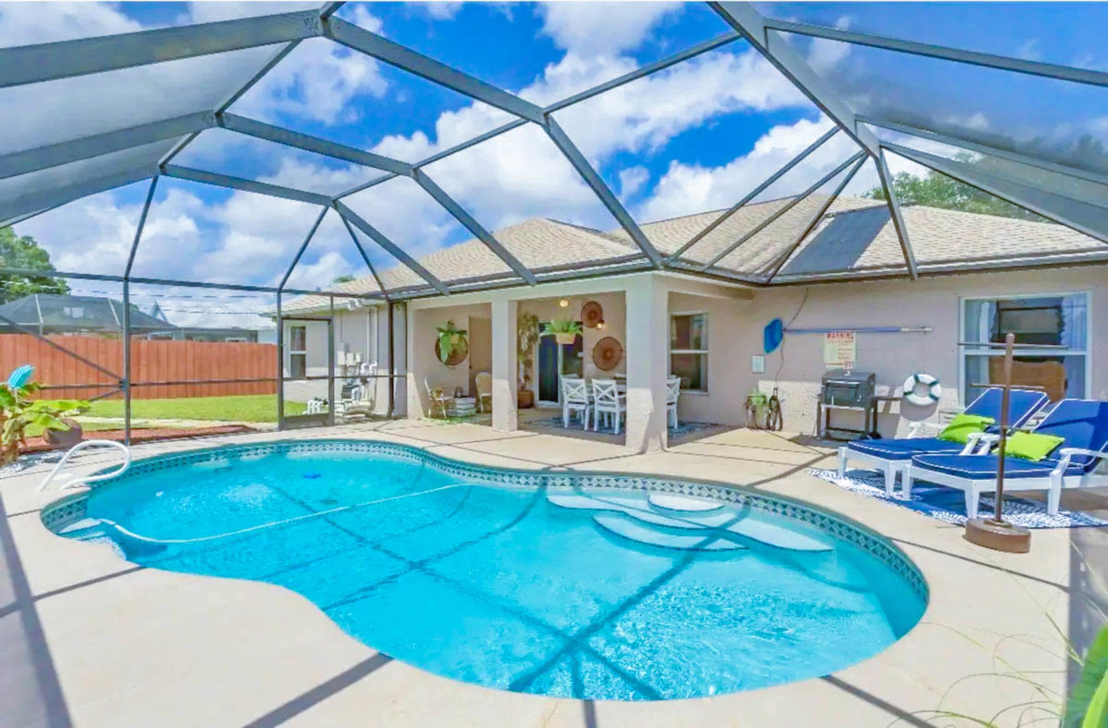 The image shows a clear, screened-in pool area with a circular swimming pool, sun loungers, and a patio space, surrounded by a well-maintained yard.
