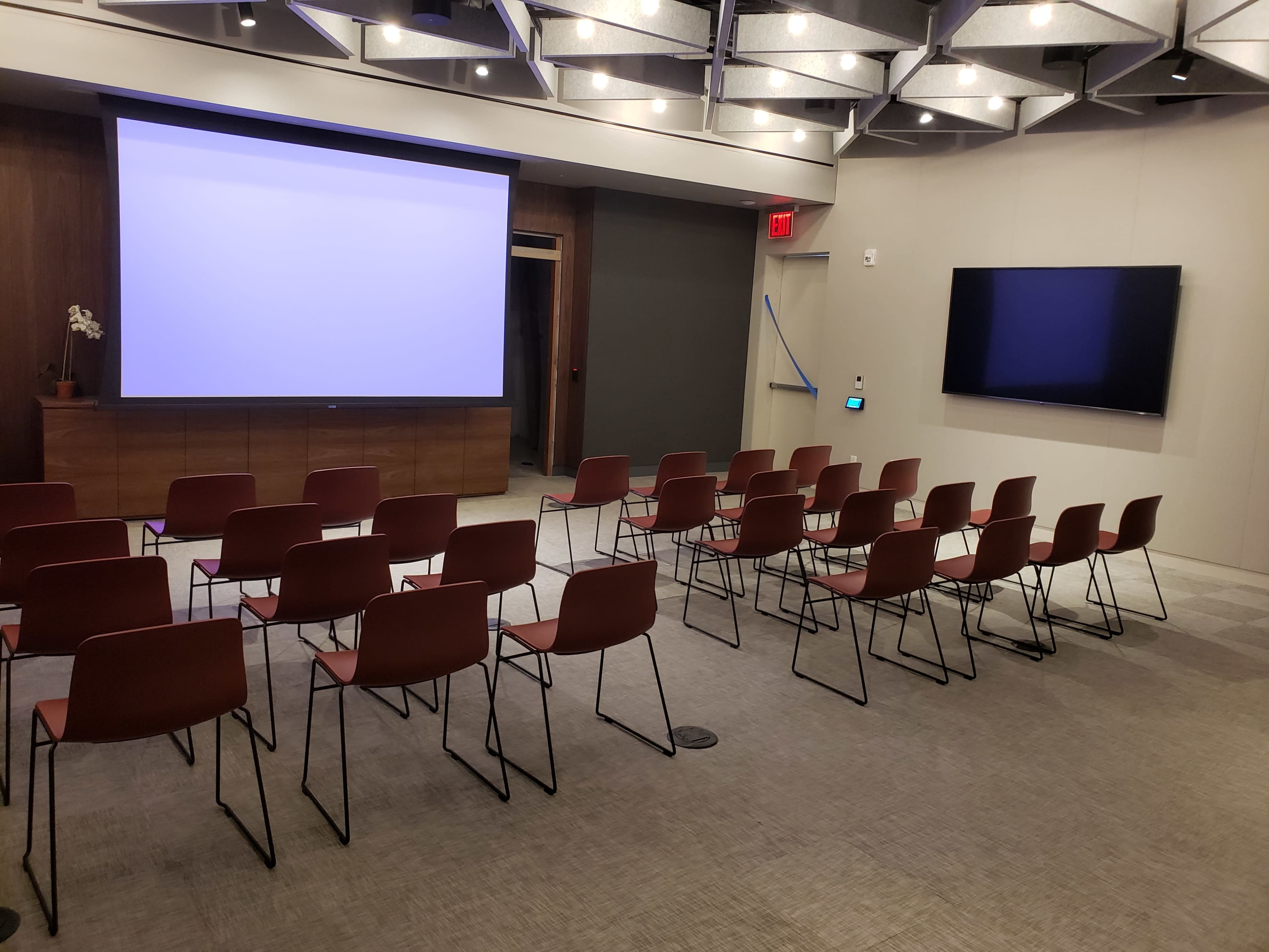 A conference room with a large projection screen at the front, rows of metal-framed chairs arranged facing it, and a flat-screen monitor on the side wall.