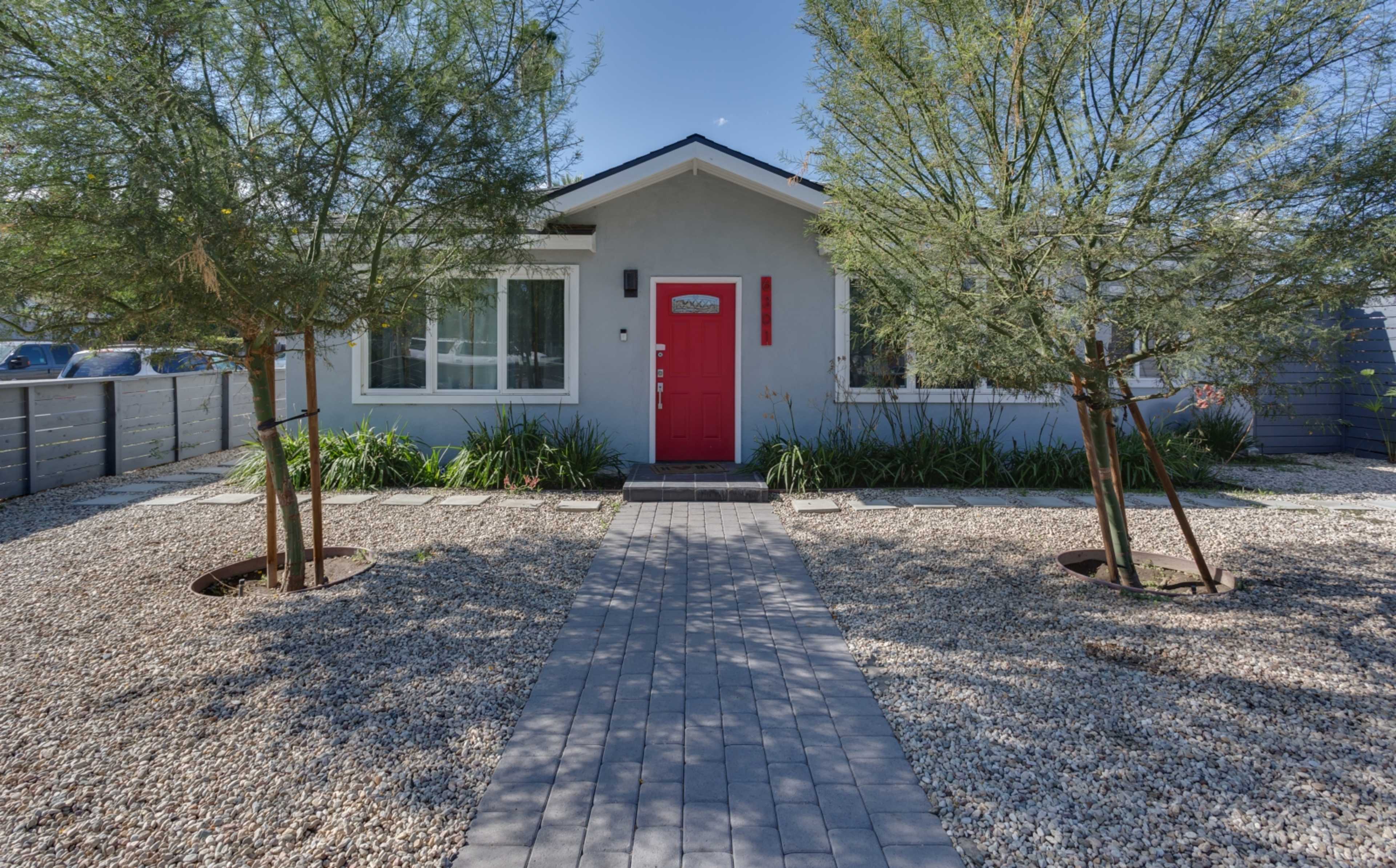 A single-story house with a vibrant red door, flanked by two trees, surrounded by gravel landscaping and a paved walkway leading to the entrance.