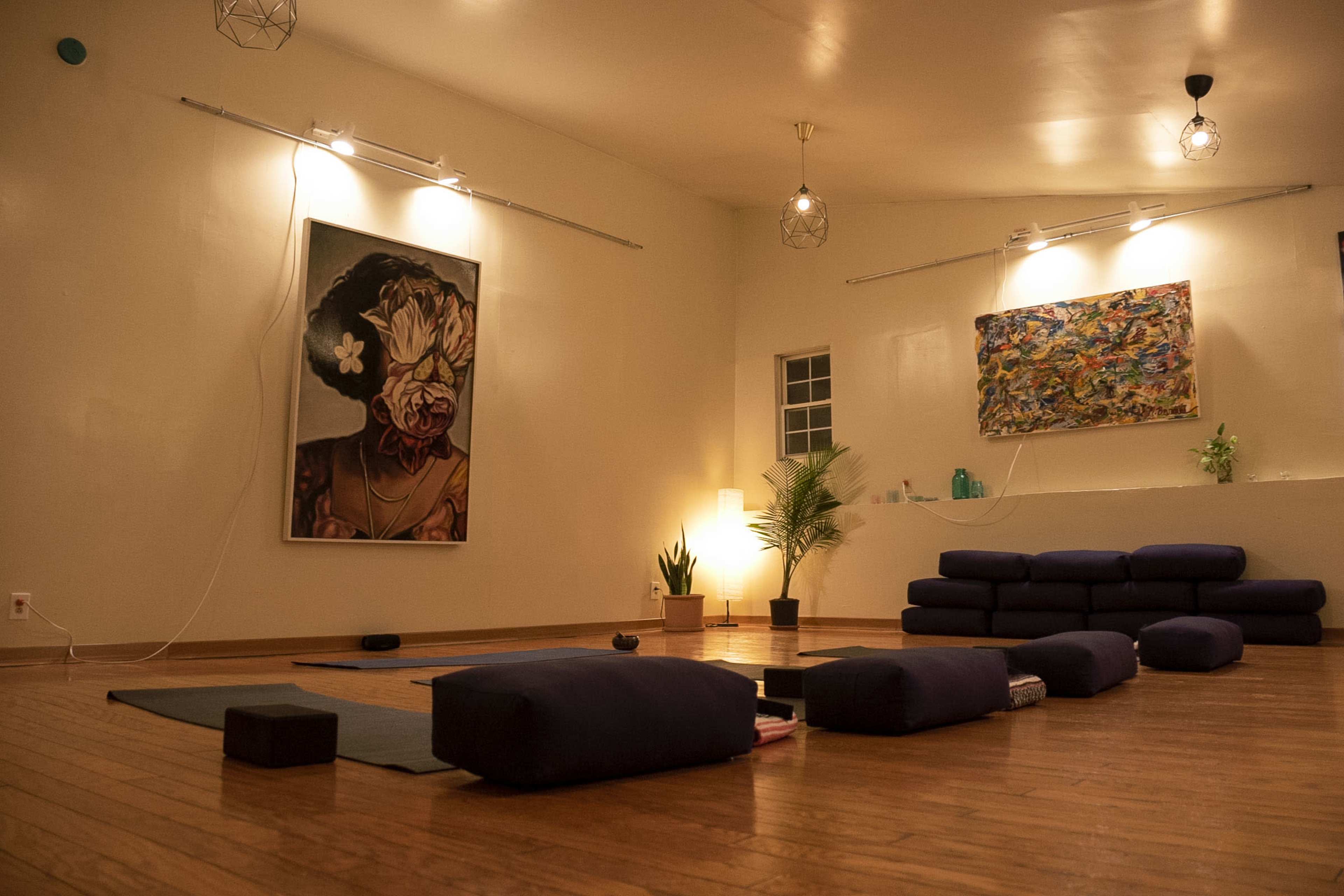 The image shows a dimly lit yoga studio with blue floor cushions, a large artwork on the wall, and a few plants in the corner.