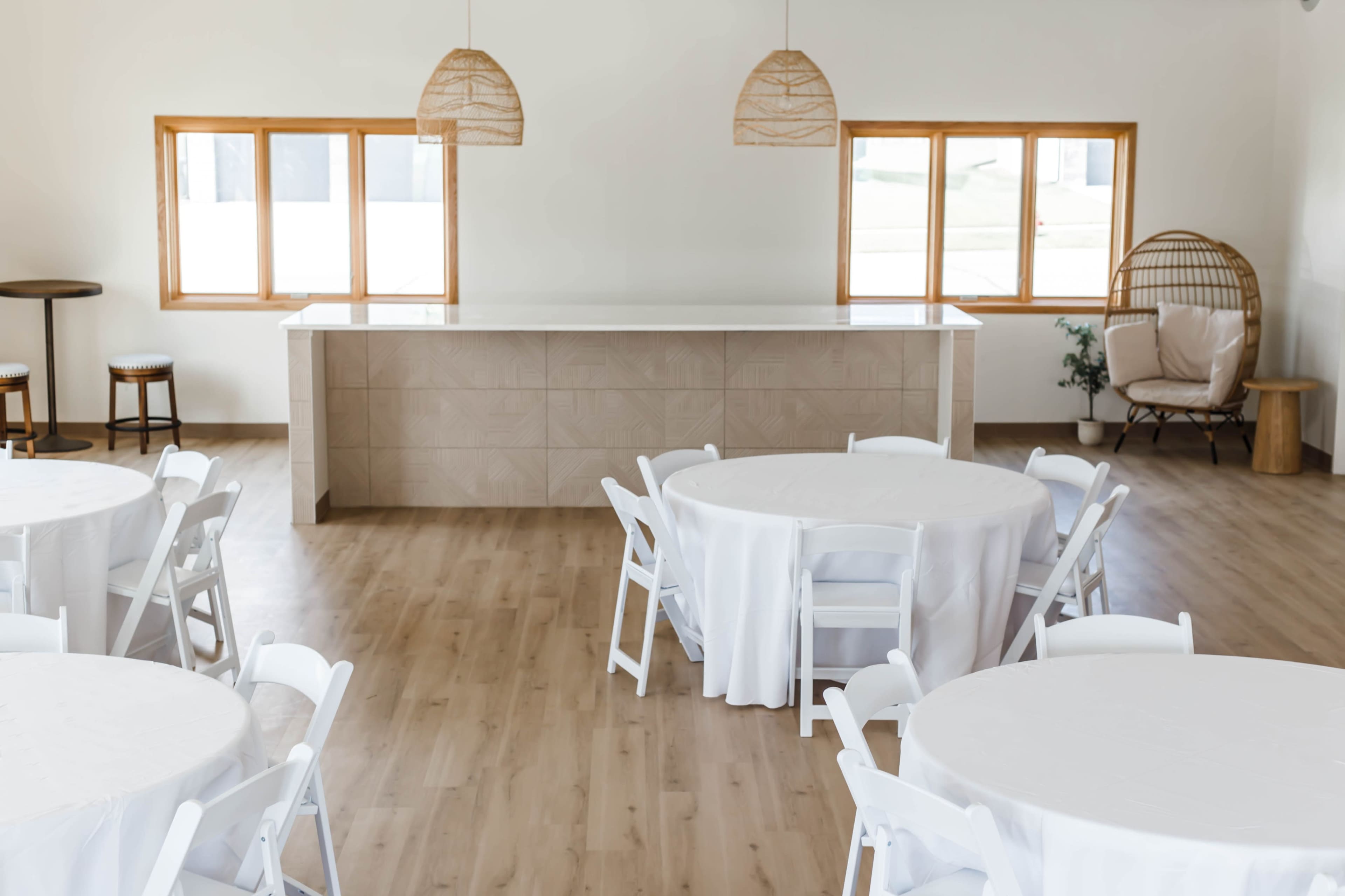 A spacious indoor event venue with round tables covered in white tablecloths, a light-colored bar area in the background, and large windows allowing natural light to enter.
