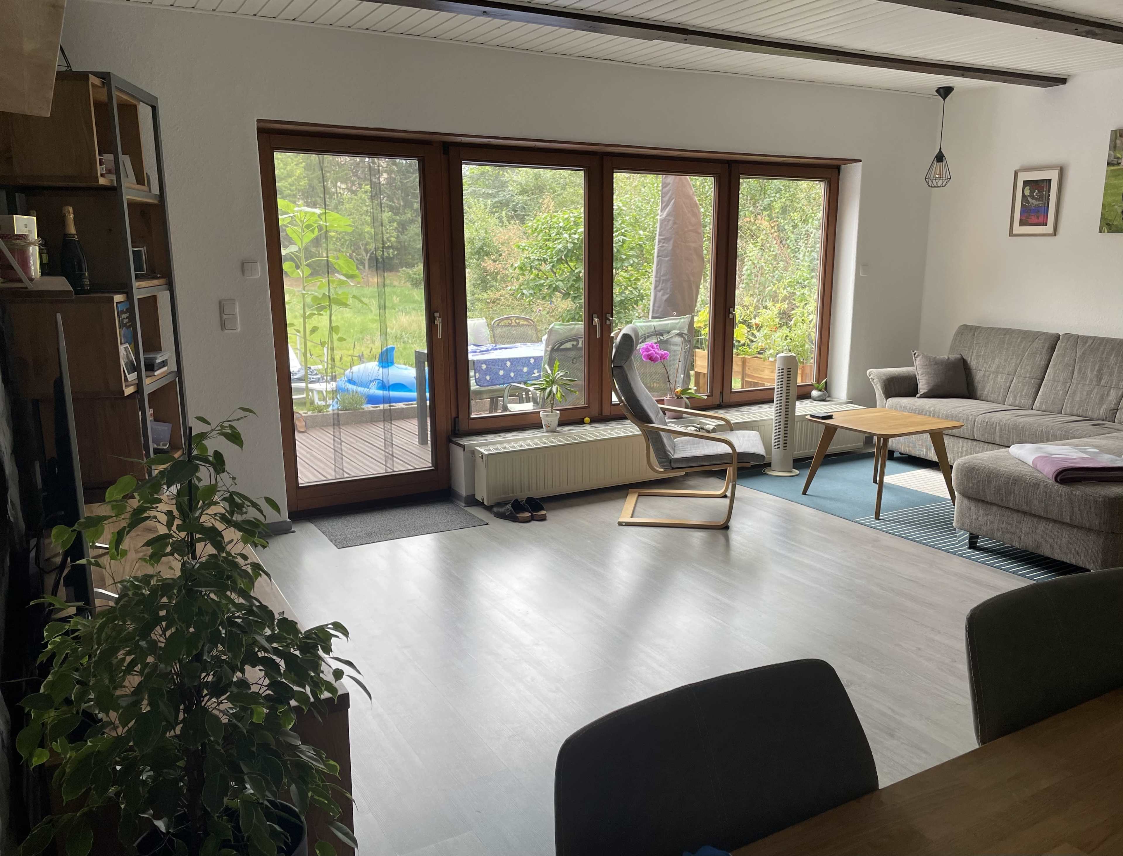 A living room with large windows, a couch, a wooden table, and a view of a garden outside.