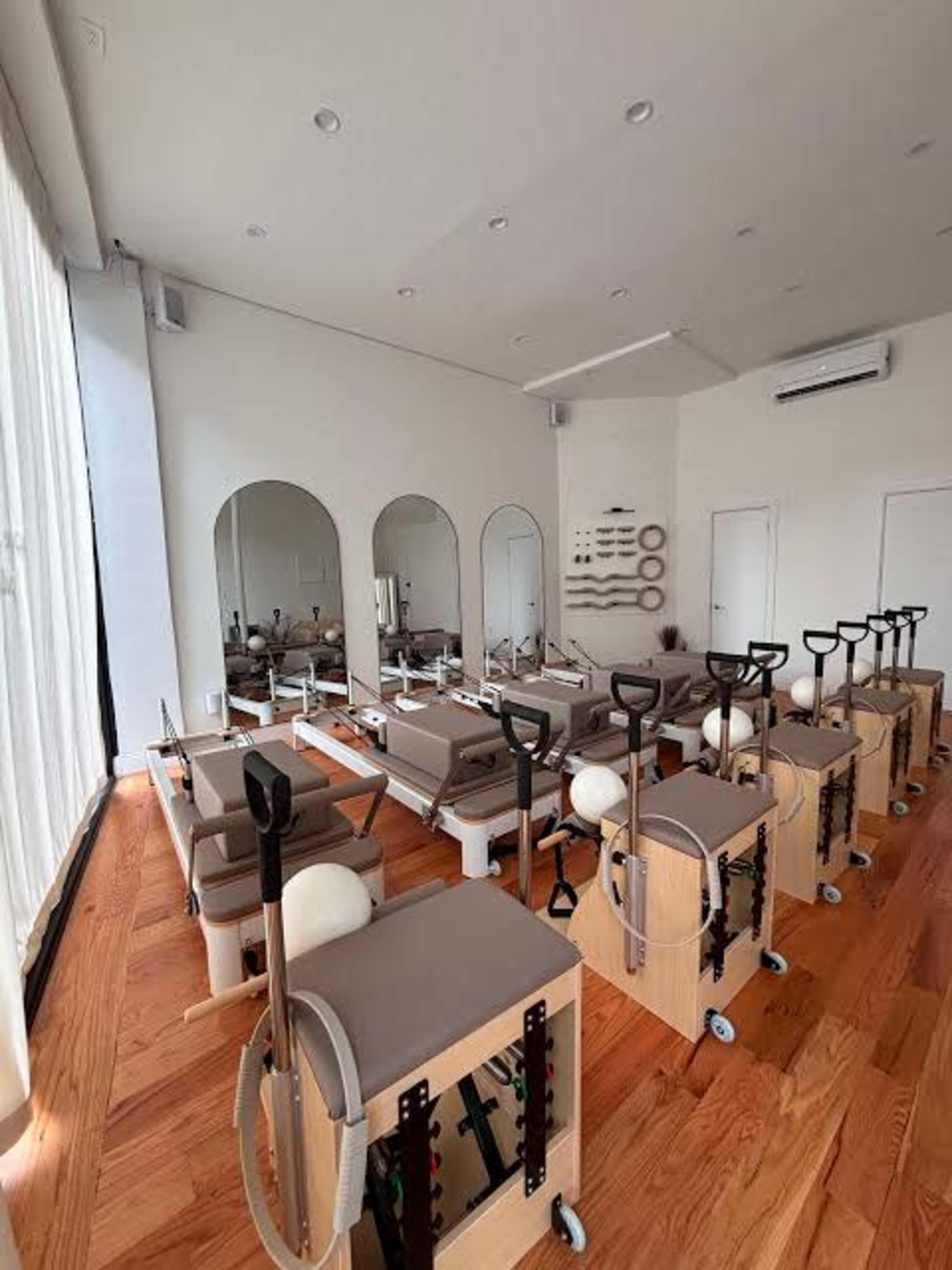 The image shows a Pilates studio with multiple reformer machines arranged neatly in rows, surrounded by mirrors and large windows.