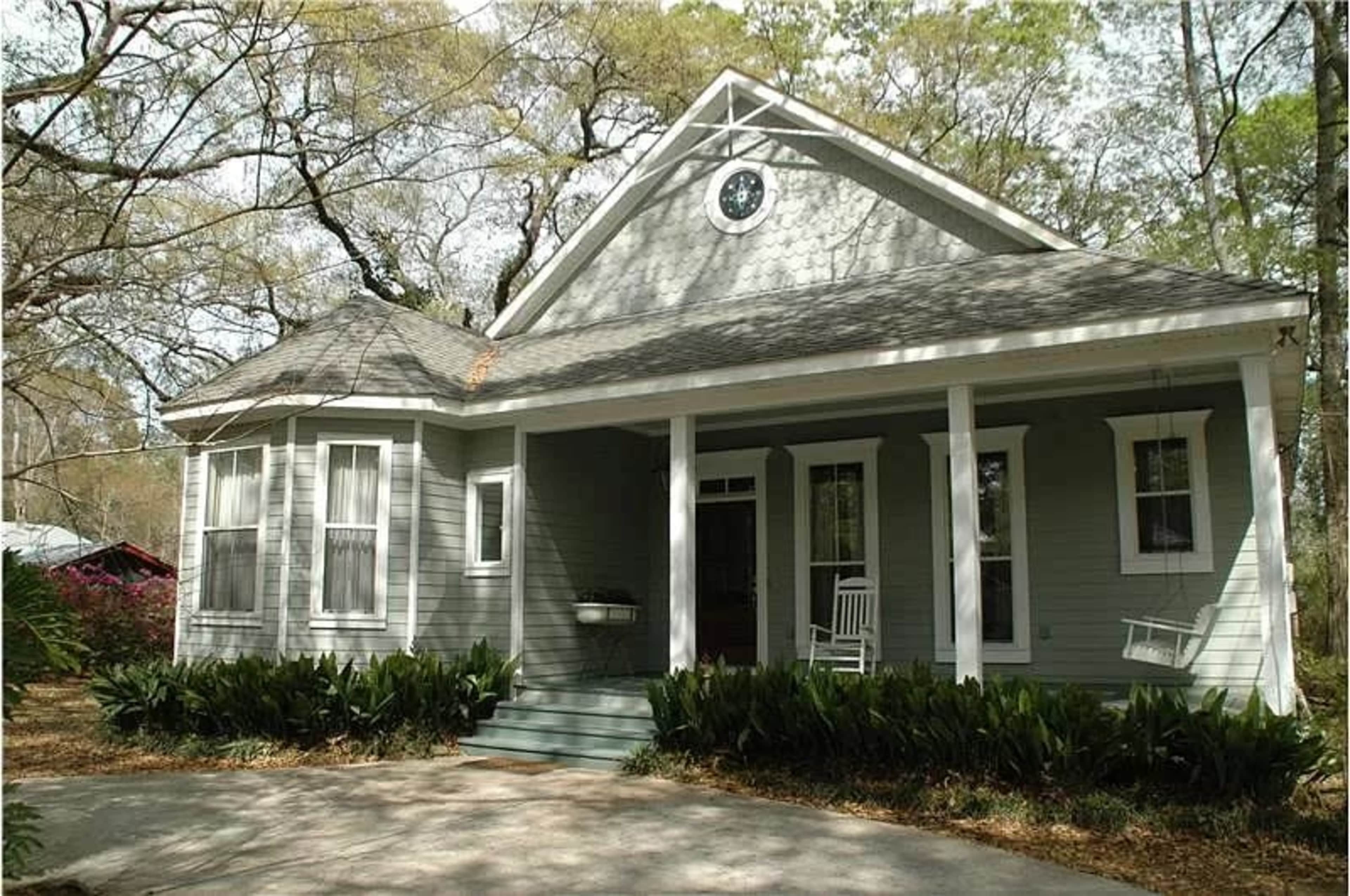 A pale green house with a gabled roof, front porch, and white rocking chairs on either side of the entrance, surrounded by greenery and bare trees.