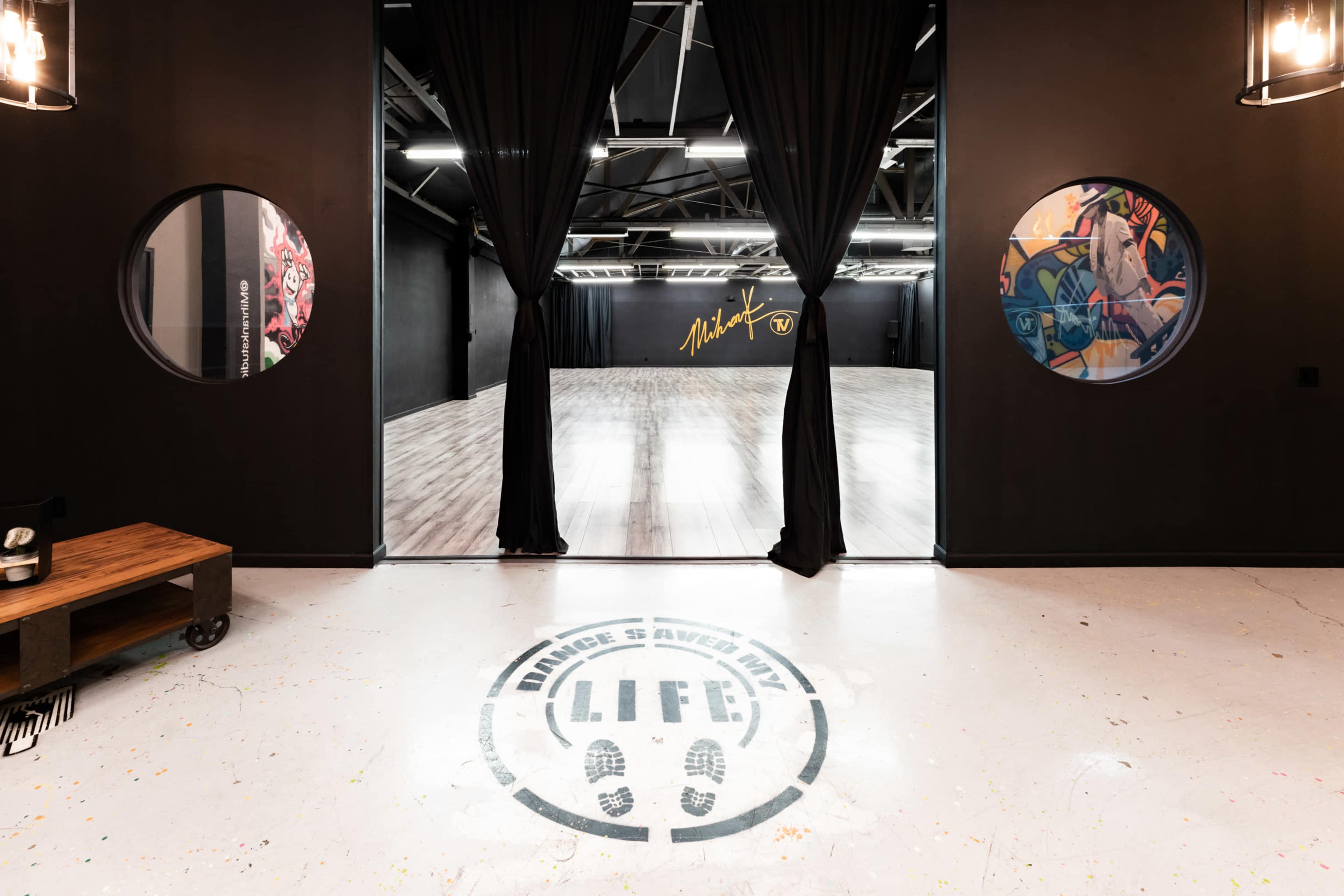 The image shows an entrance to a dance studio featuring round windows, a dark interior with curtains, and a space marked with wooden flooring and colorful wall art.