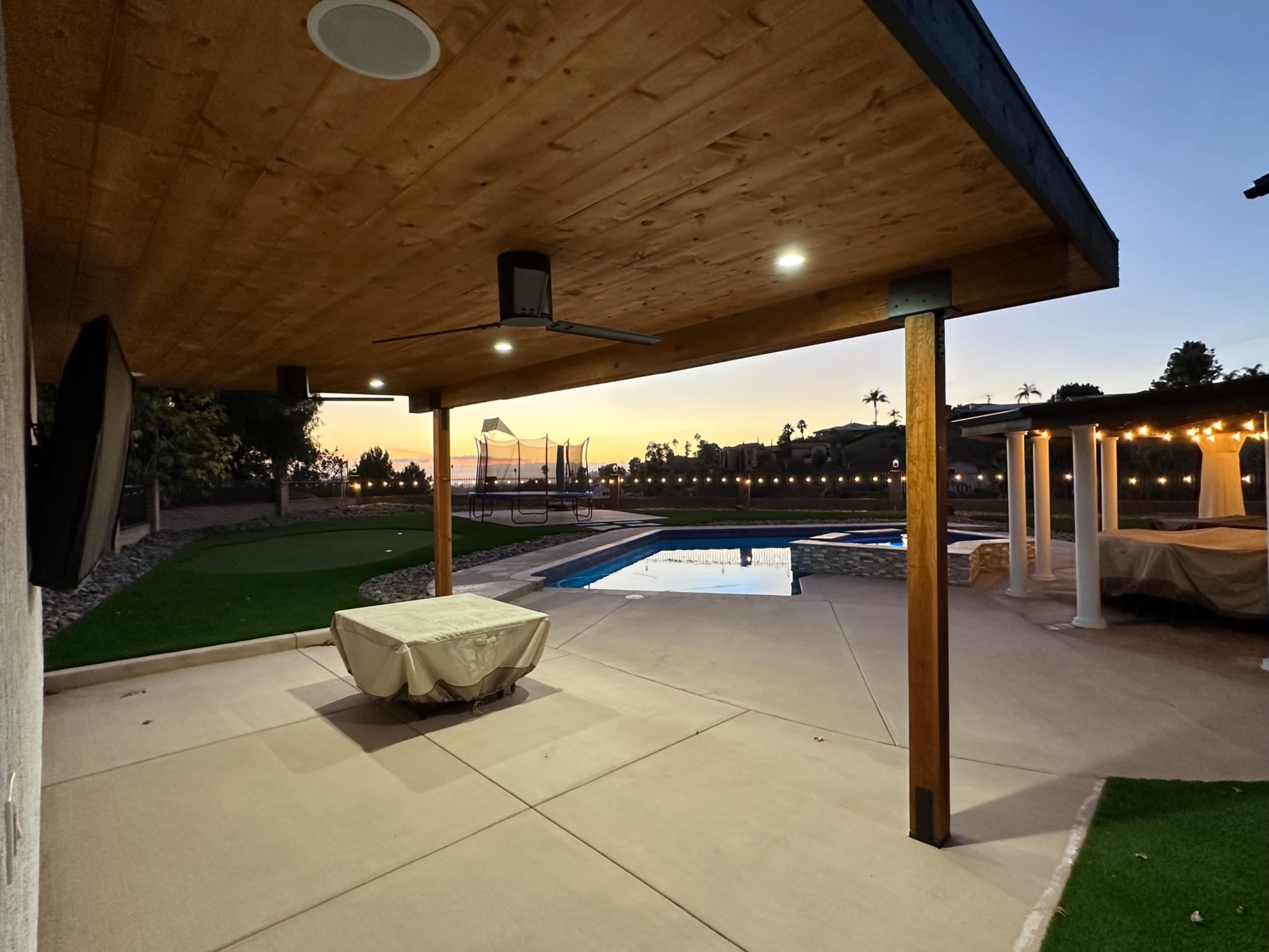 A covered patio with a ceiling fan, overlooking a pool area at sunset, surrounded by lawns and a trampoline in the background.