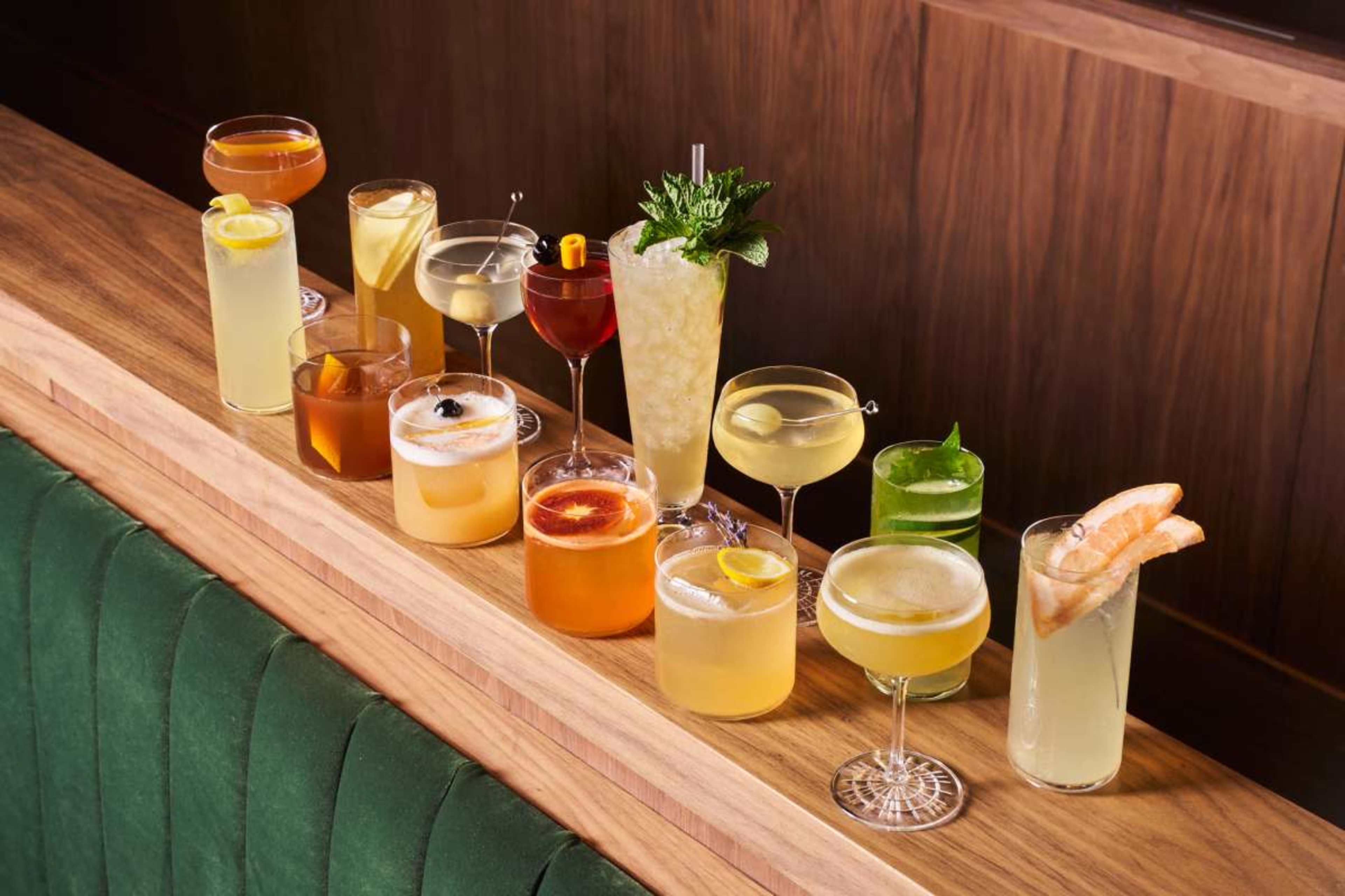 The image shows a selection of twelve different cocktails arranged on a wooden bar top, featuring various garnishes and glass styles.
