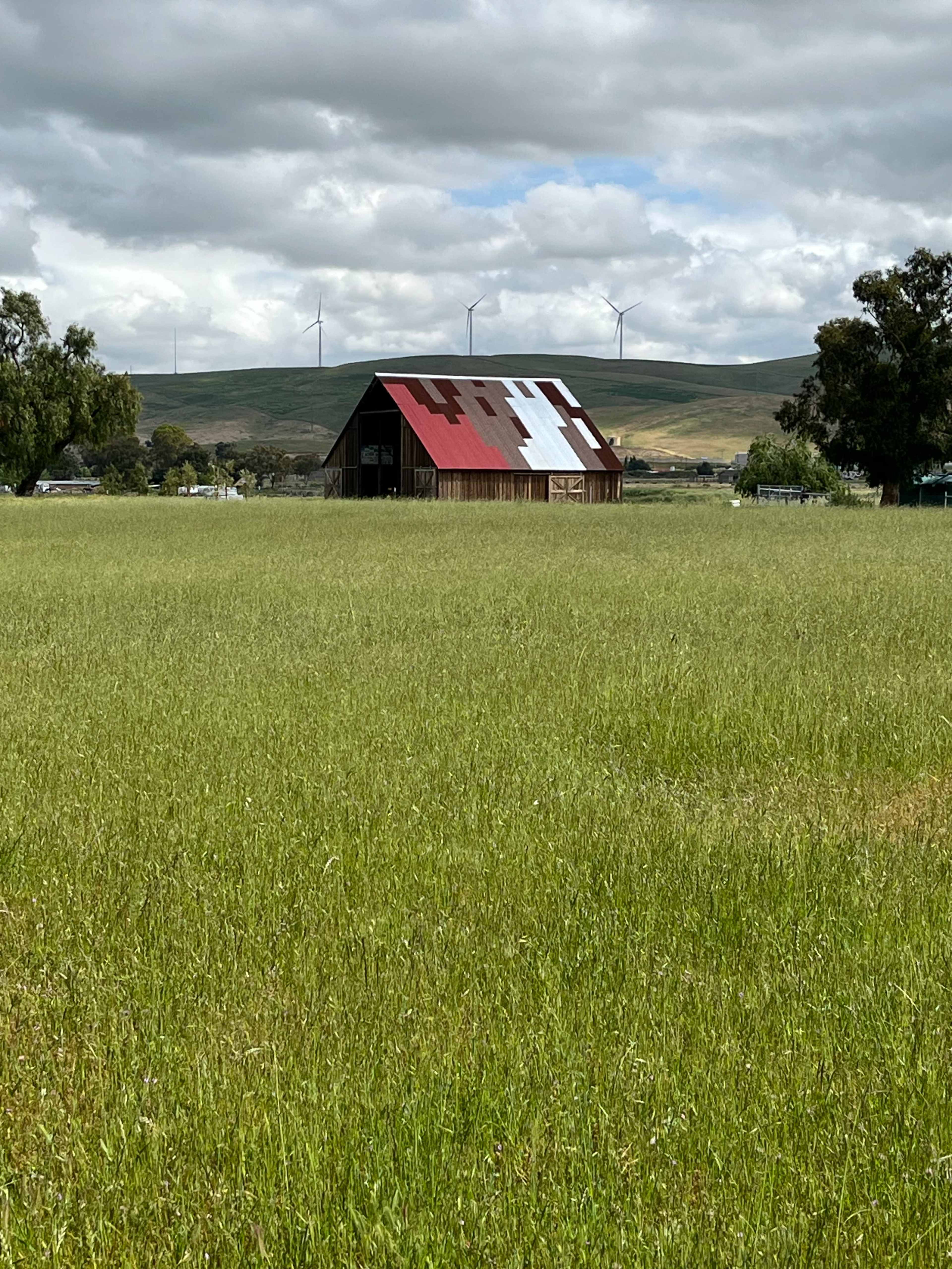 A weathered barn with a red and white roof stands in a grassy field, framed by distant wind turbines and rolling hills under a cloudy sky.