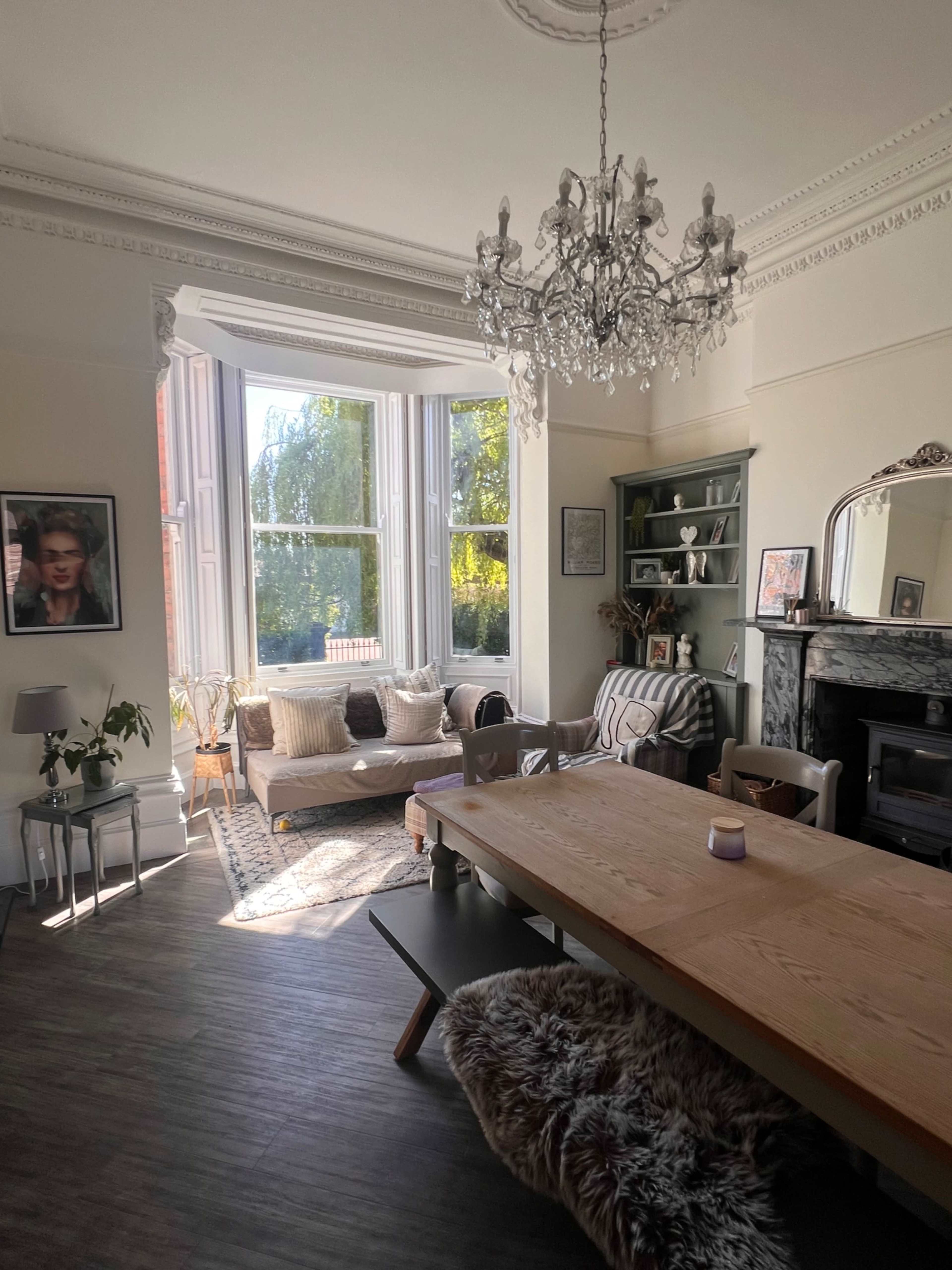 The image shows a well-lit dining area in a stylish room featuring a large wooden table, a plush sofa, a chandelier, and a decorative bookshelf.