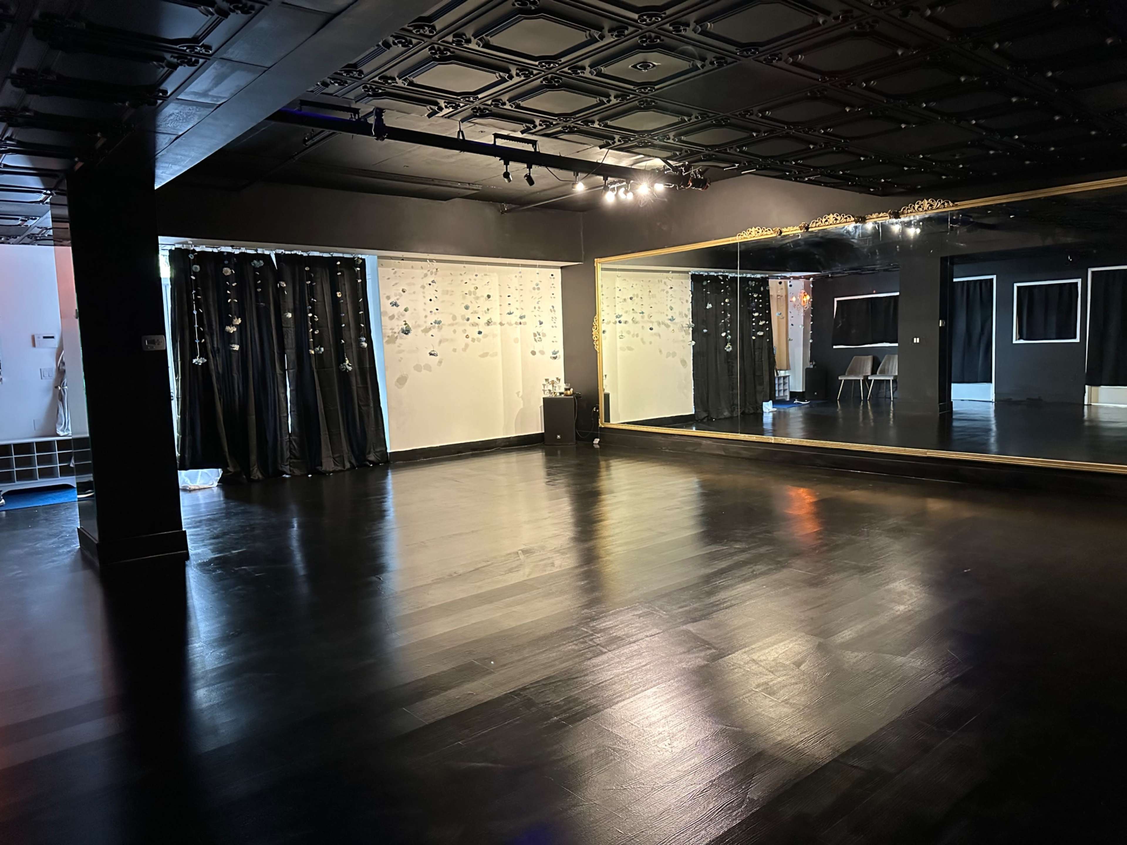 An empty dance studio features a large mirrored wall, black wooden flooring, and sheer curtains adorned with hanging decorations.