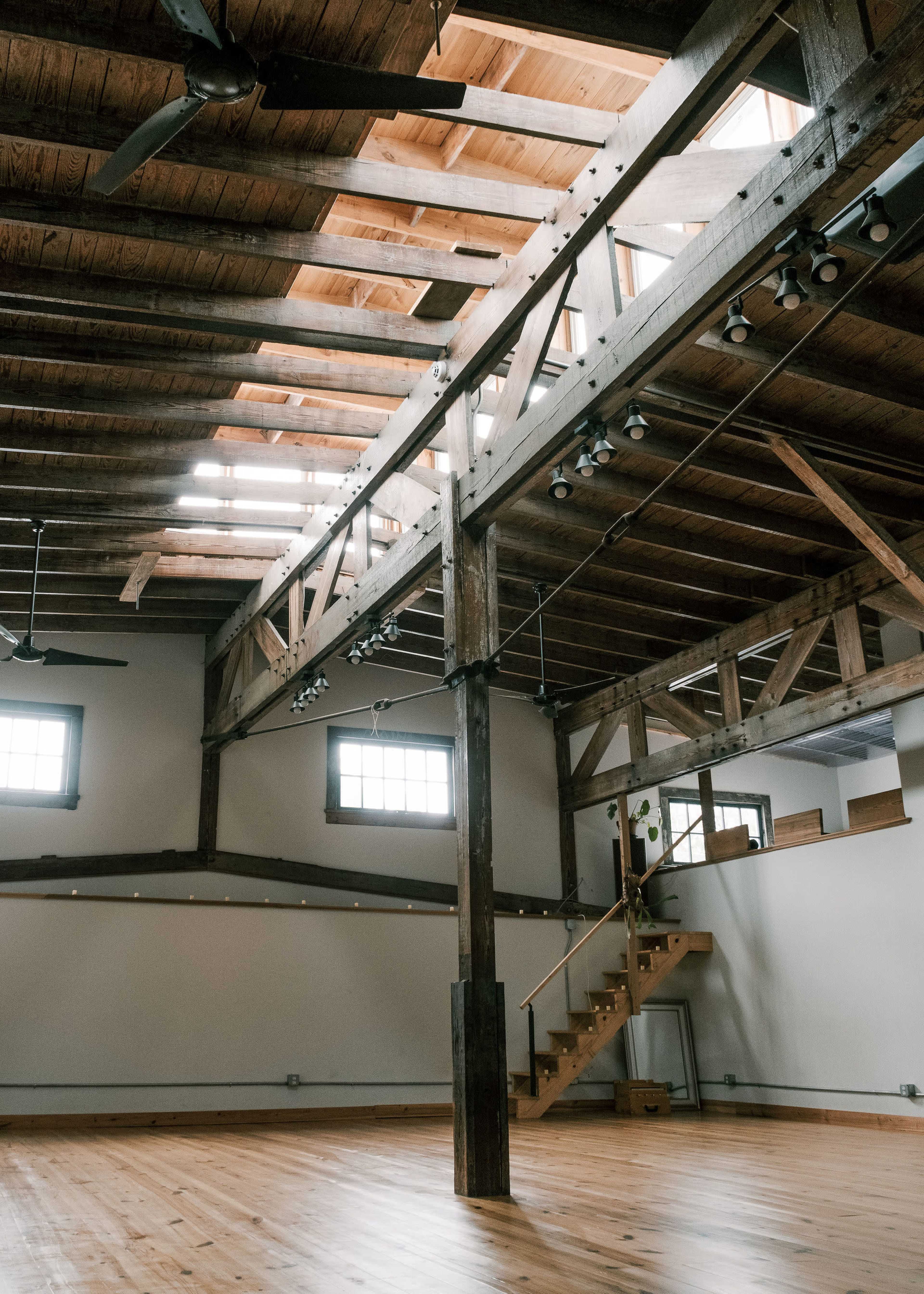 The image shows a spacious interior with wooden beams, high ceilings, and a wooden staircase leading to a second level.