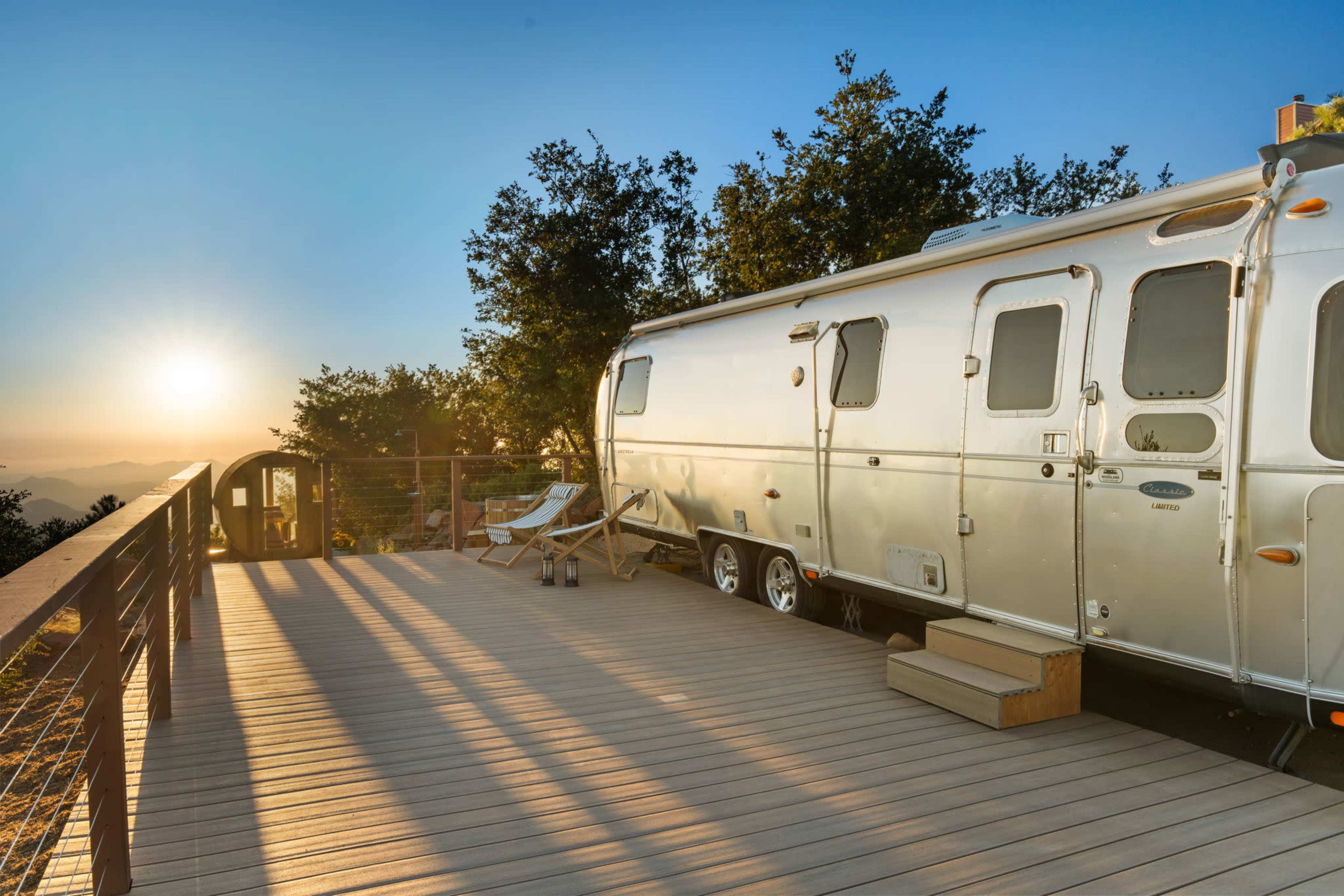 A shiny Airstream trailer is parked on a wooden deck with sunlight streaming through trees and mountains in the background.