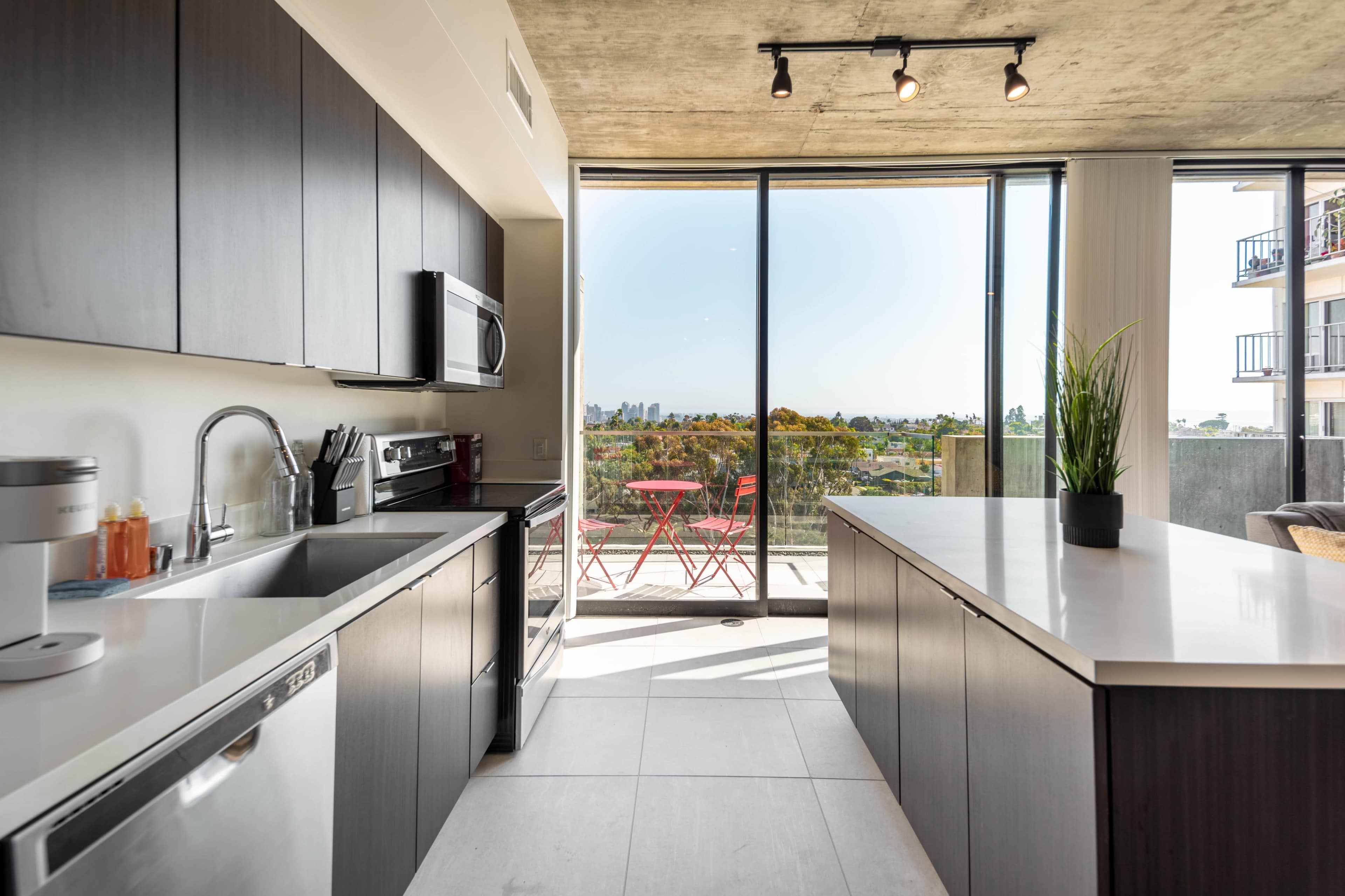 A modern kitchen features stainless steel appliances, dark cabinetry, and large windows that overlook a balcony with a view of the city and surrounding trees.