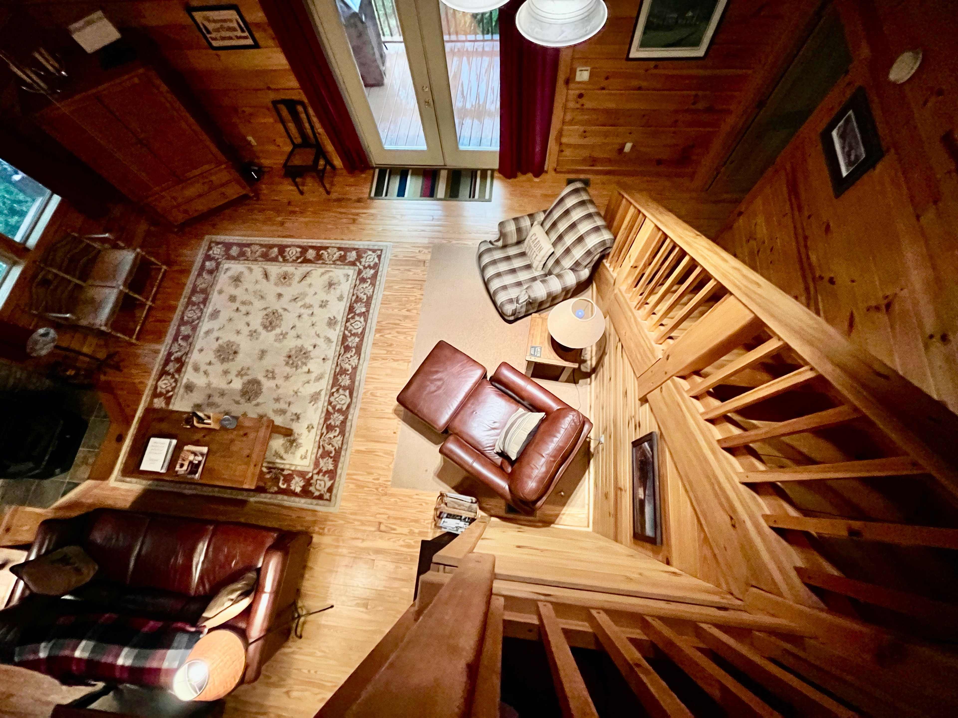 An overhead view of a wooden-furnished living room with a staircase, a leather recliner, a patterned rug, and sliding glass doors leading outside.