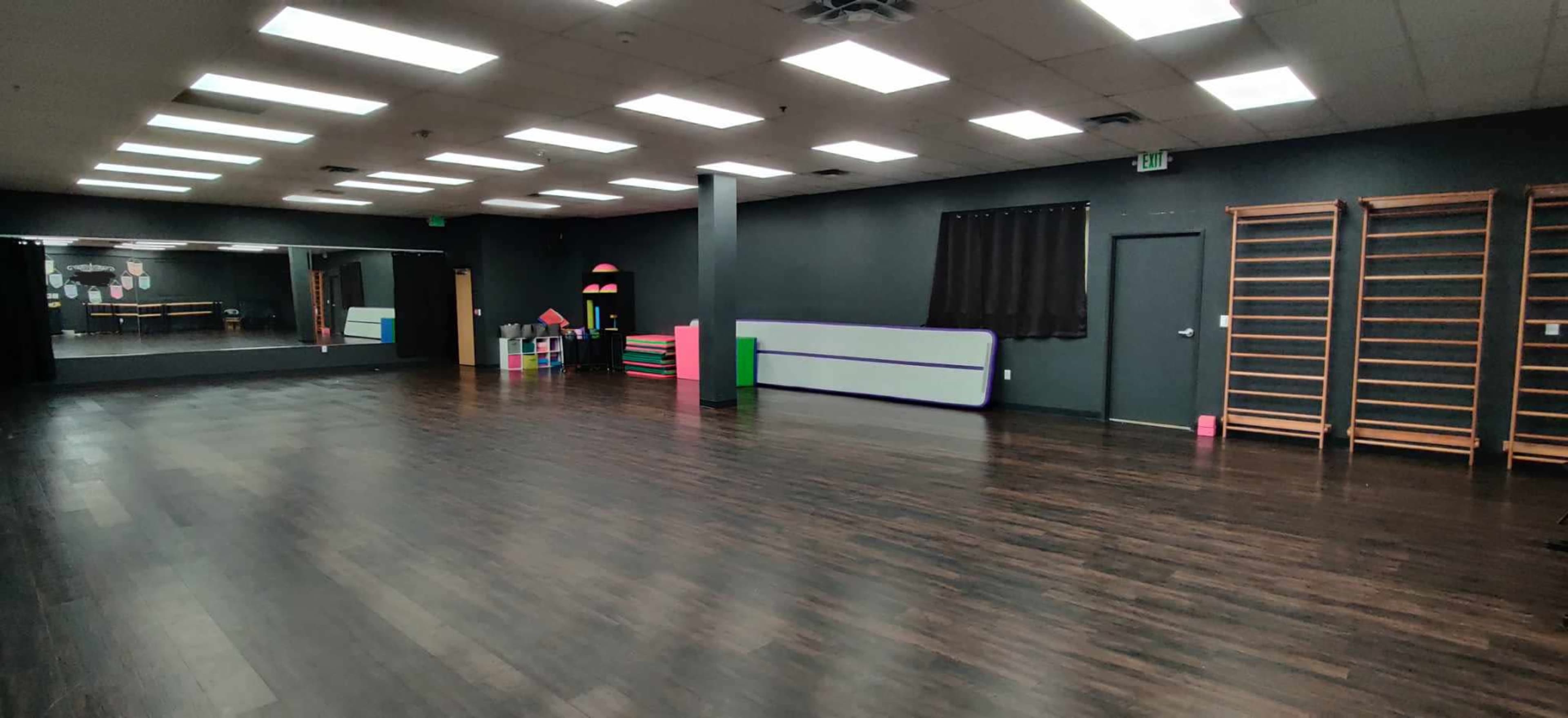 An empty indoor studio with dark walls and wooden flooring, featuring a large mirror, exercise equipment, and a black curtain.