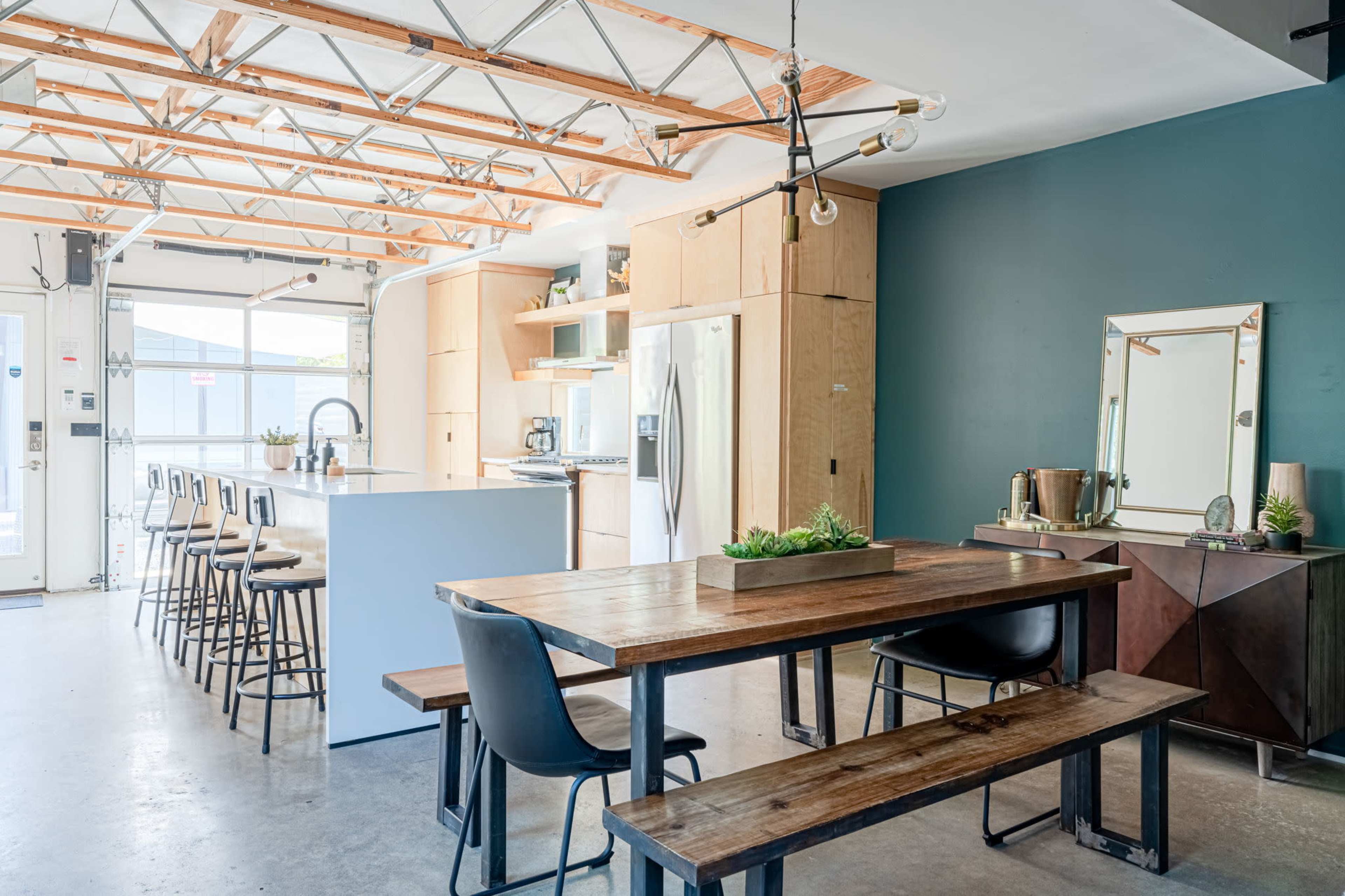 The image shows a modern kitchen and dining area featuring a large wooden dining table, high stools at a kitchen island, and minimalist cabinetry with a dark accent wall.