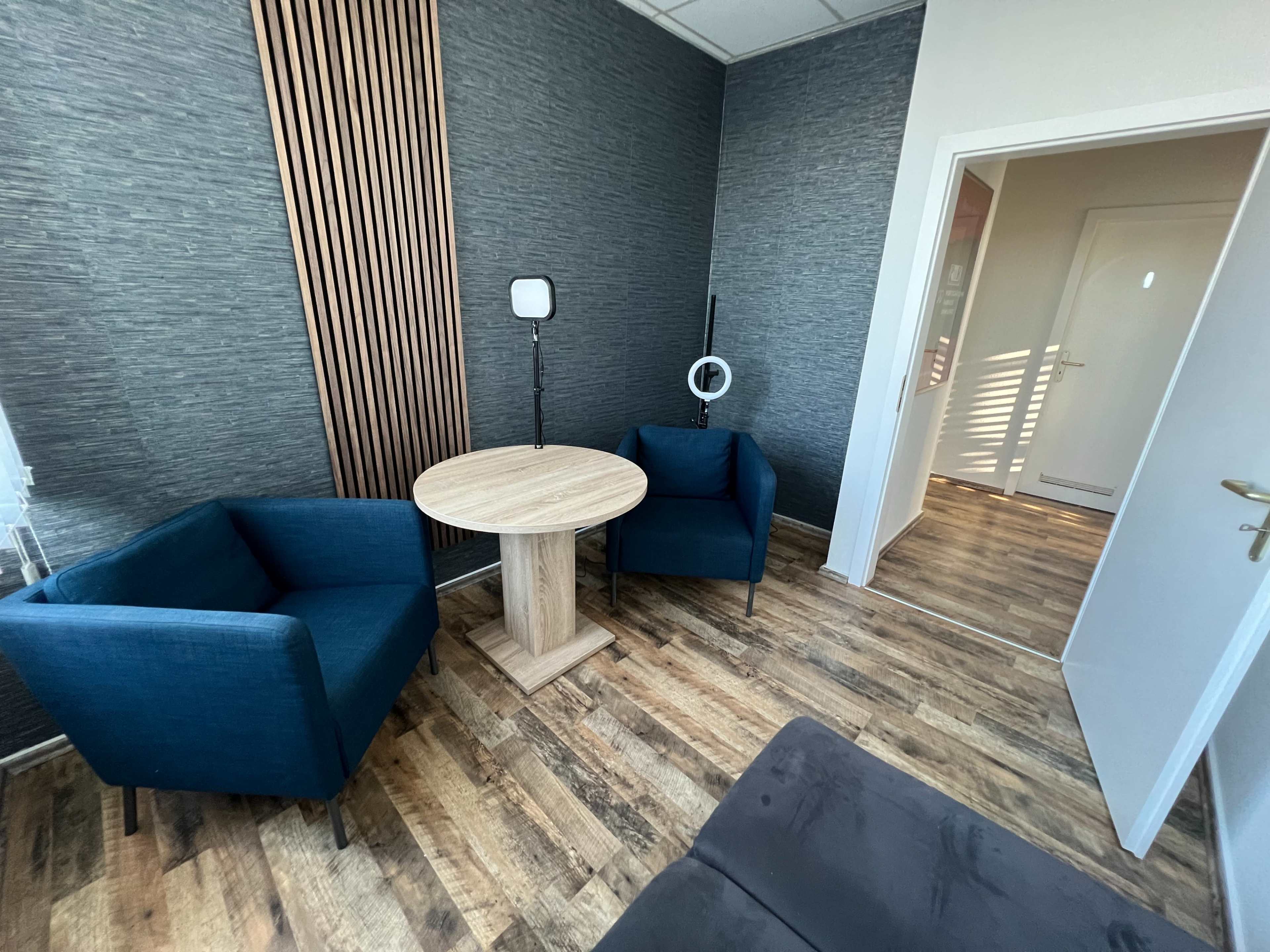 The image shows a small, modern waiting room with two blue chairs, a round table, and a doorway leading to another room.