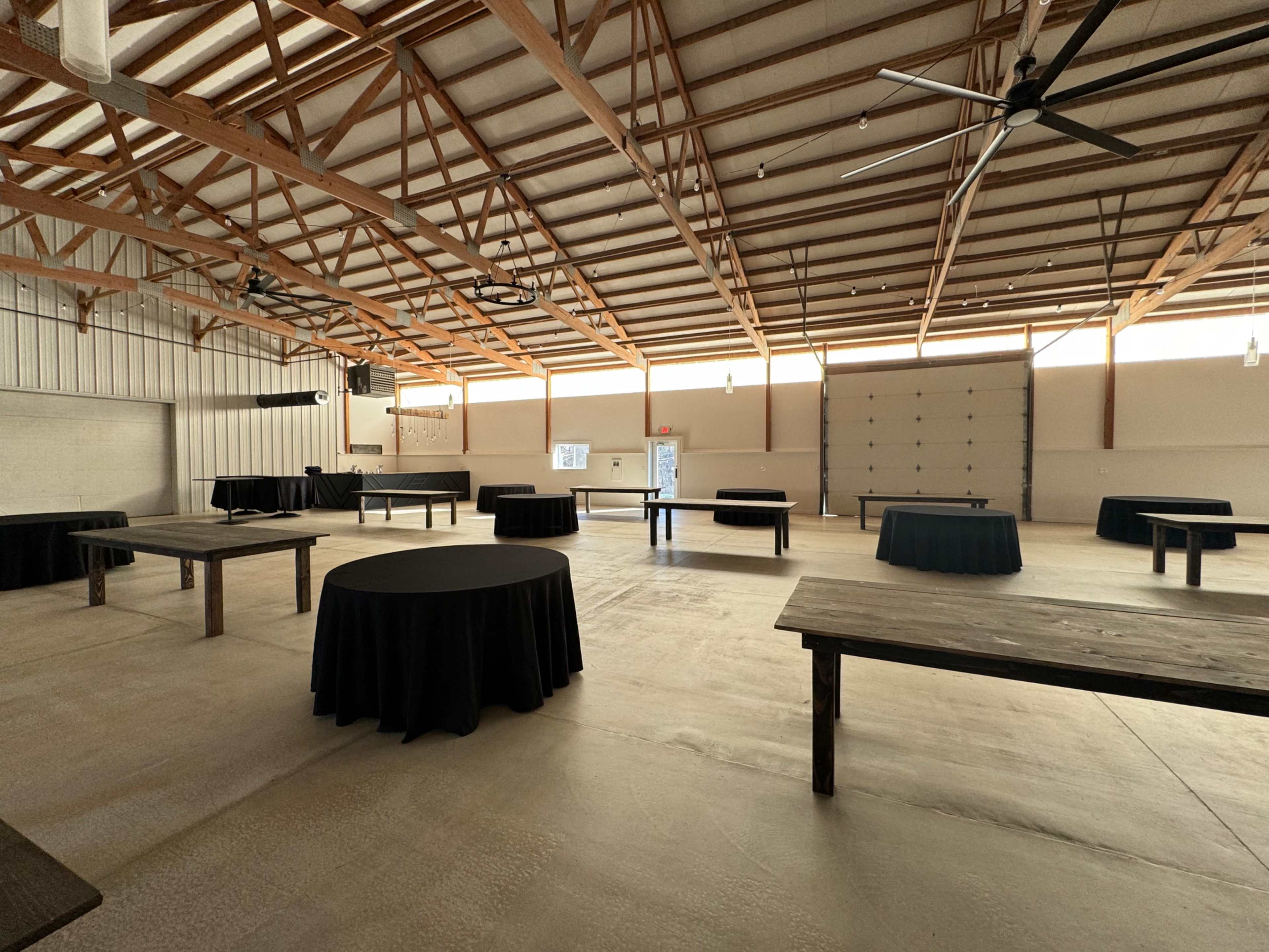 The image shows an empty indoor event space featuring several round tables covered with black tablecloths beneath a high wooden ceiling with exposed beams.