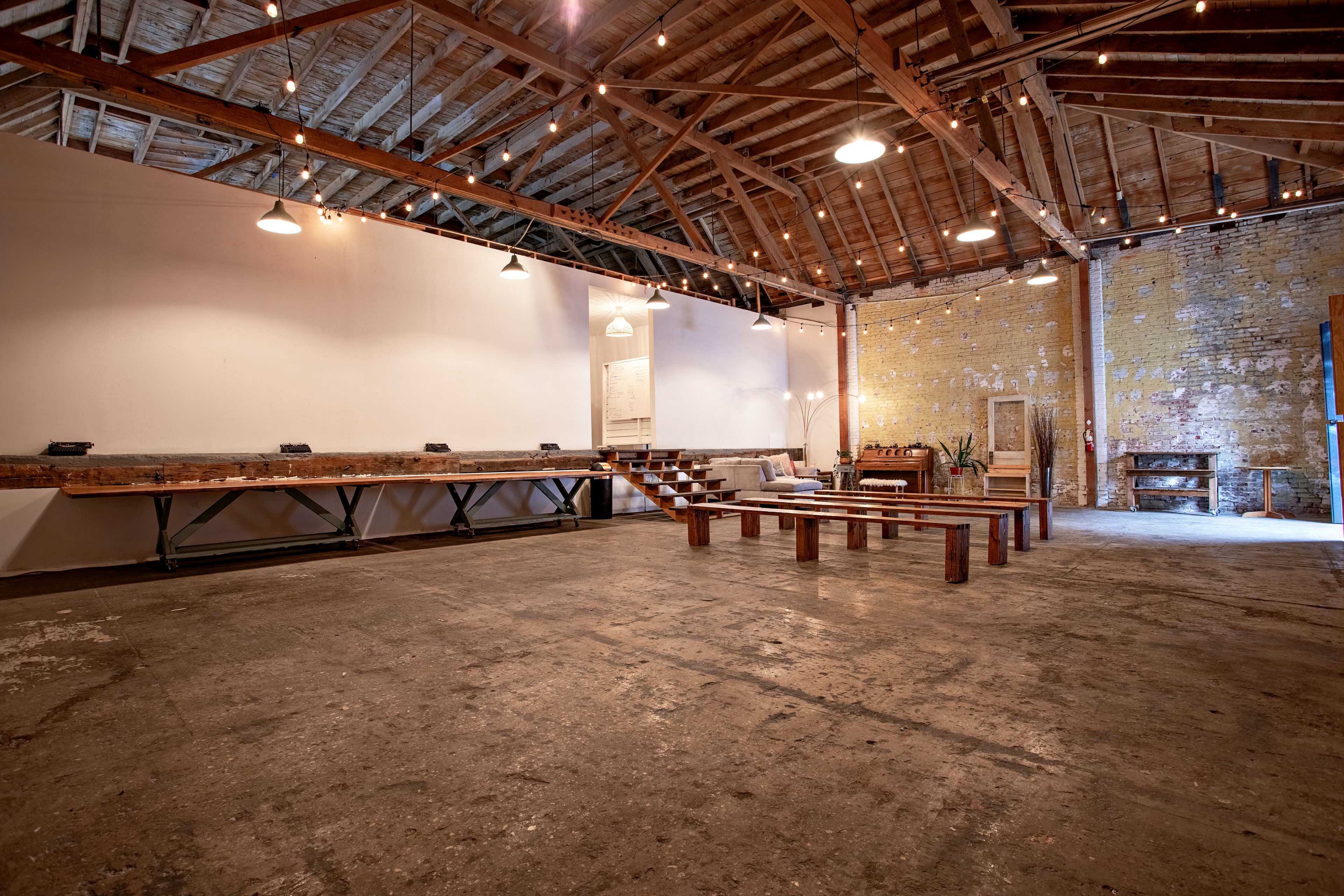 An empty industrial-style venue with exposed wooden beams, a polished concrete floor, and simple seating arrangements.