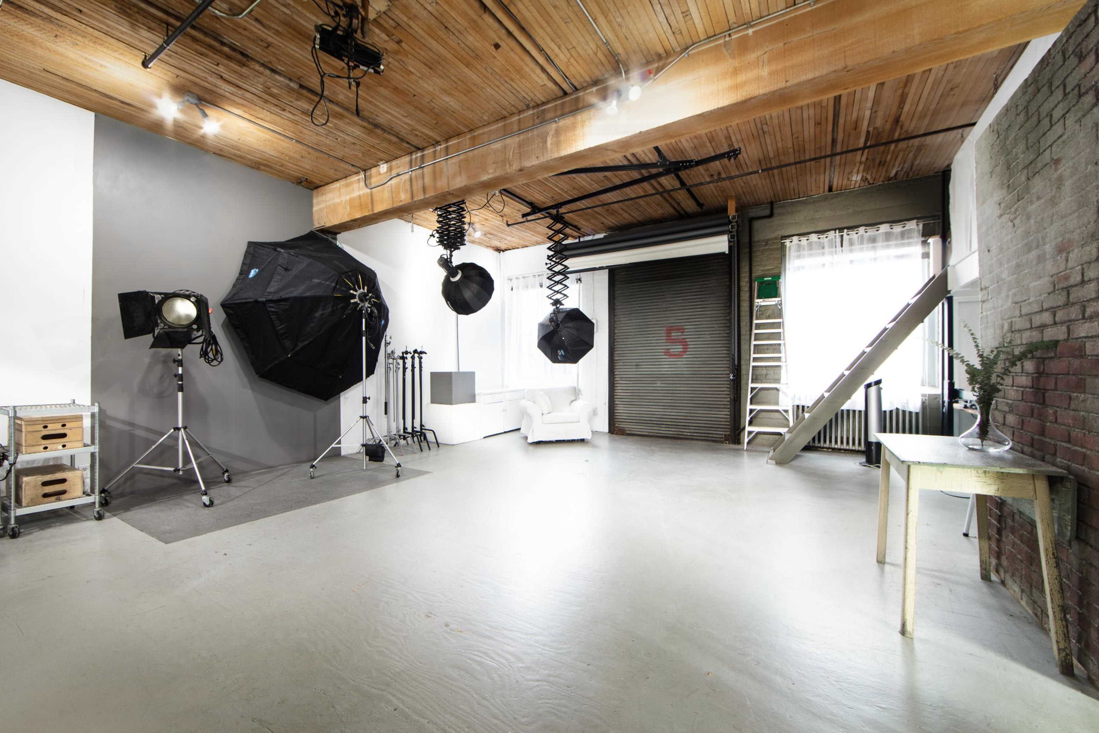 A spacious photography studio with various lighting equipment, a white couch, and industrial-style elements such as exposed wooden beams and a metal garage door.