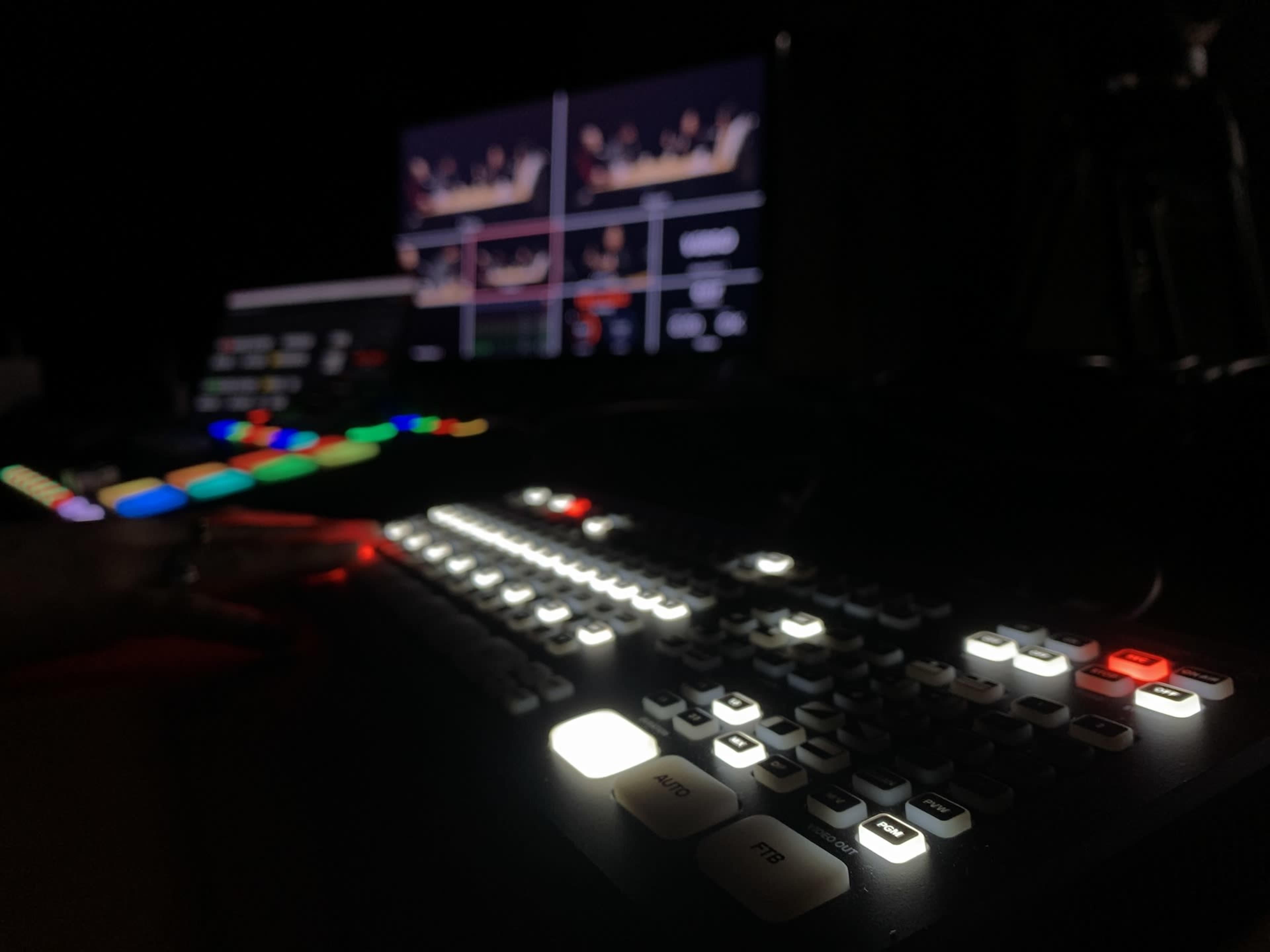 The image shows a dimly lit control panel with illuminated buttons and screens displaying video feeds.