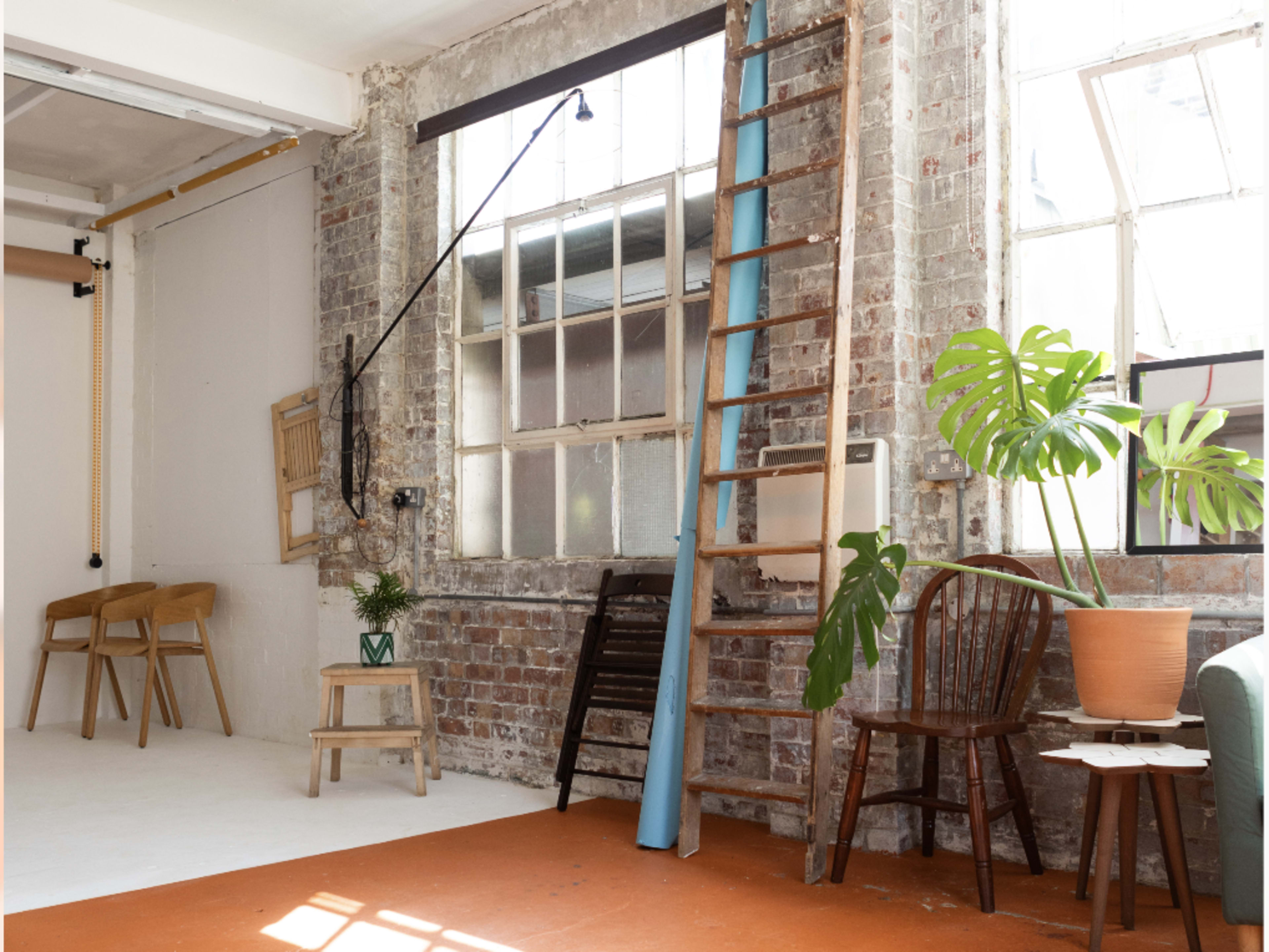 The scene features a well-lit studio space with exposed brick walls, a wooden ladder leaning against a wall, various chairs, a potted plant, and a blue backdrop rolled up beside a window.