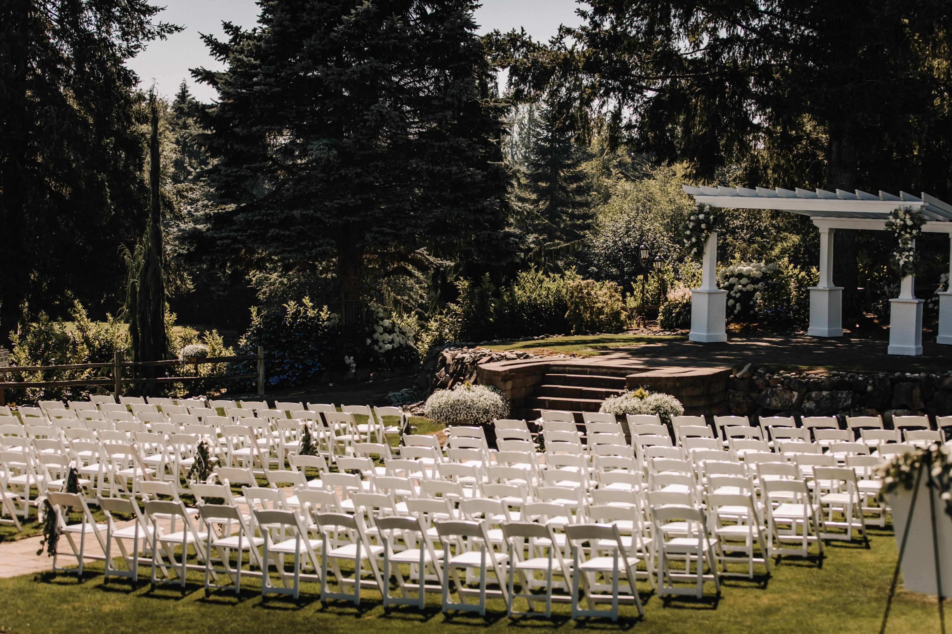 A large outdoor wedding venue features rows of white chairs arranged in front of a decorated arch on a sunny day.
